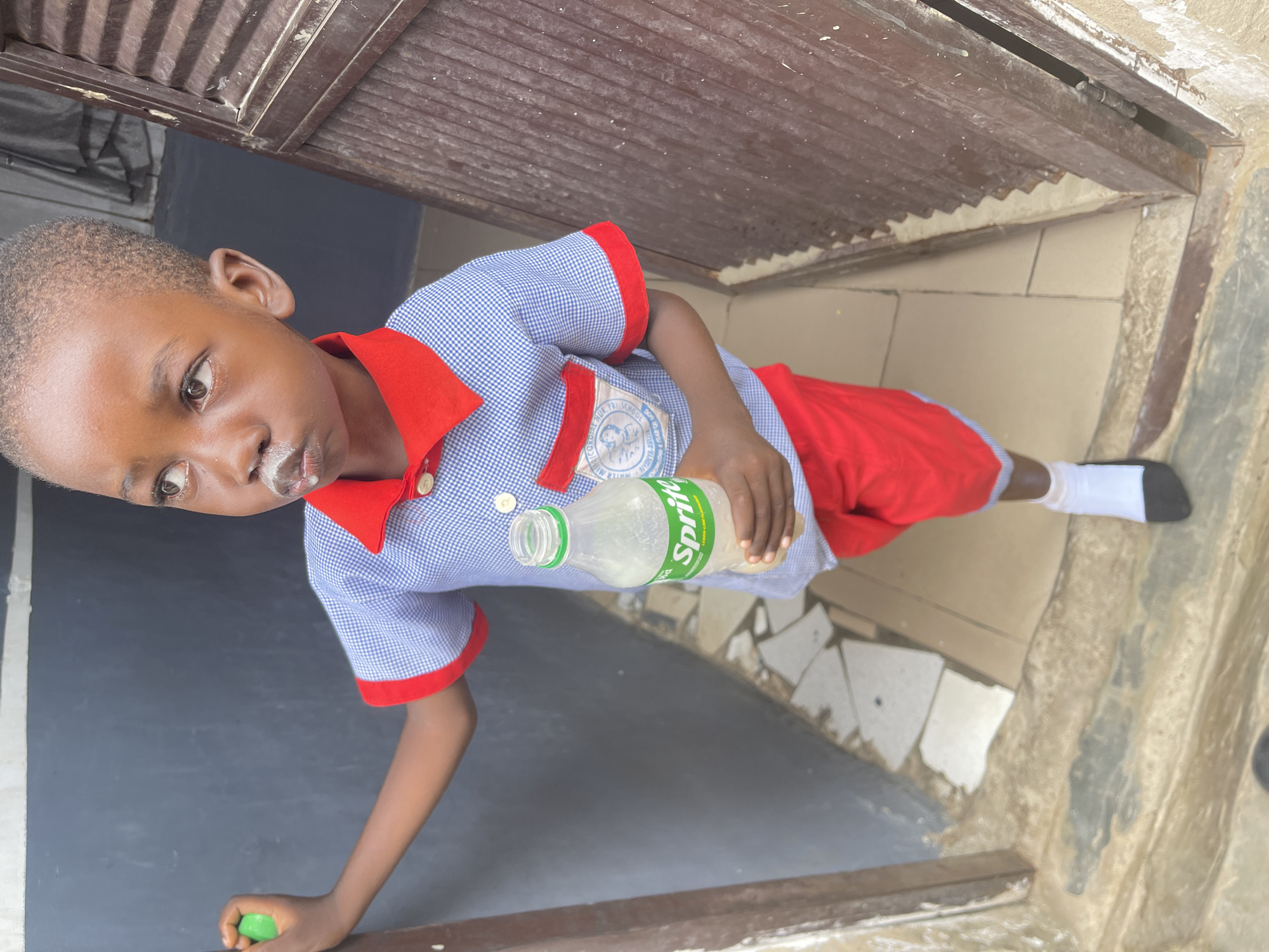 A young child, likely a boy, stands in what appears to be a doorway or entrance area in Nyanya, Nigeria. The child is wearing a blue and white checkered short-sleeved school uniform shirt featuring a red collar and red cuffs, complemented by red shorts. A patch on the shirt is partially visible, showing text such as "NUR-PAL SCHOOL" and "MERRY NURSERY". White residue, possibly from a milky or creamy drink, is present on the child's upper lip. In their right hand, the child holds a plastic bottle of Sprite, clearly displaying its green logo. A small, bright green object, potentially a bottle cap or a toy, is held in the child's left hand. The child's gaze is directed slightly upwards and to their left, with a neutral expression. The background consists of a dark wall to the child's left, a light and dark tiled floor with an irregular pattern, and a brown, horizontally grooved structure overhead, consistent with a door frame or ceiling. The lighting suggests it is daytime. The child is also wearing white socks and dark shoes.