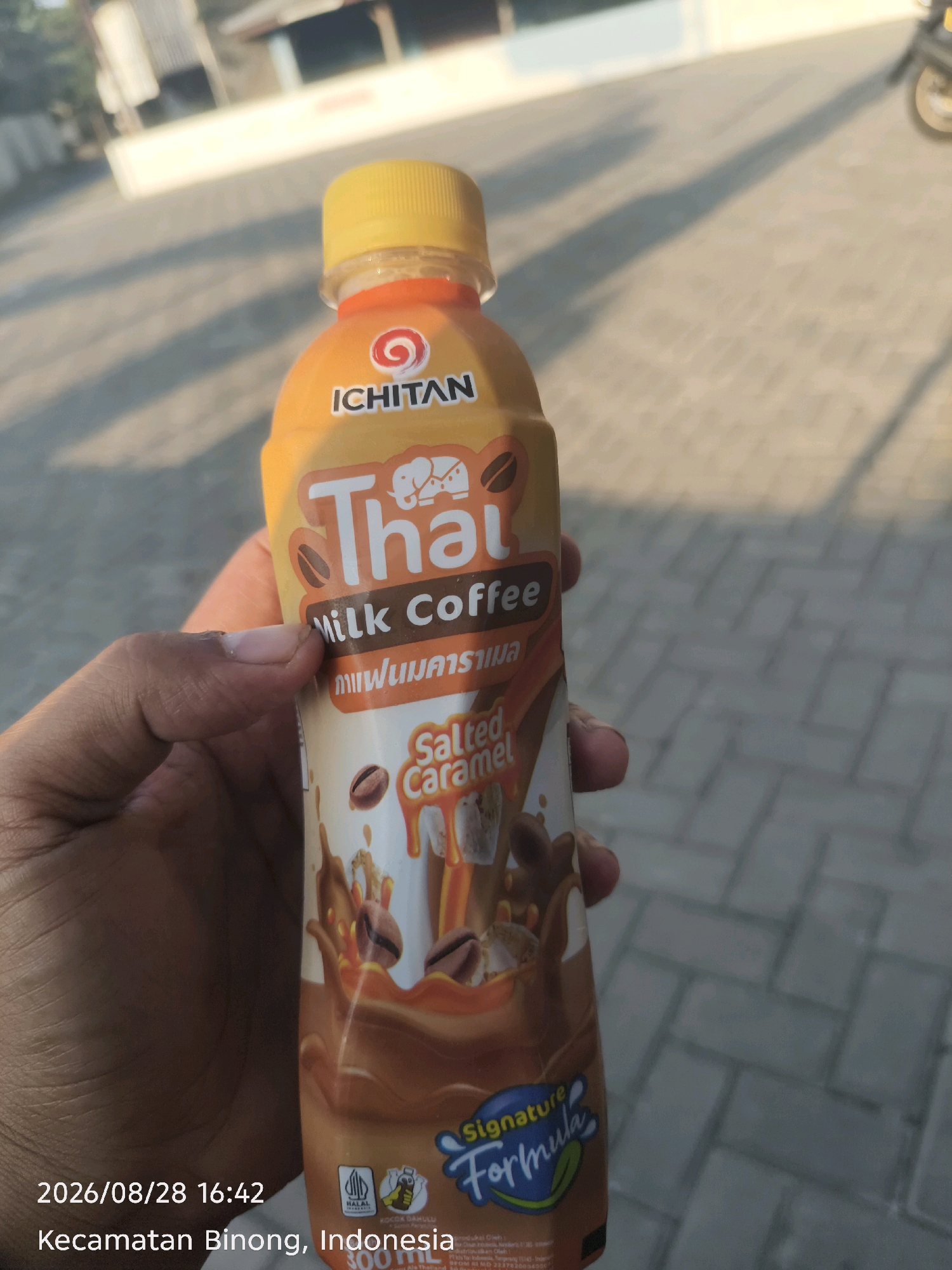 A person's hand holds a bottle of Ichitan Thai Milk Coffee, Salted Caramel flavor, outdoors. The bottle has a yellow cap and an orange and brown label featuring Thai text, an elephant graphic, and coffee beans. The background is a blurred paved area with a building and a motorcycle visible. The timestamp "2026/08/28 16:42" and the location "Kecamatan Binong, Indonesia" are overlaid on the lower left of the image.