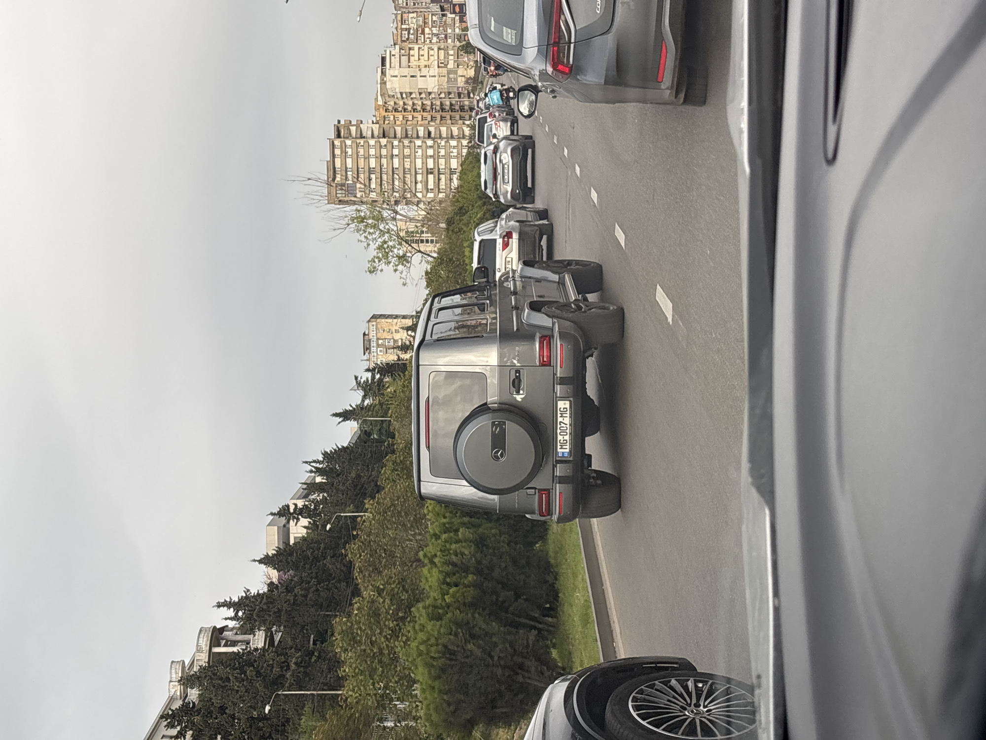 The image captures a street scene in Tbilisi, Georgia, dominated by a line of cars stuck in traffic. Prominently featured is a gray Mercedes-Benz G-Class SUV with a spare tire mounted on the rear door and a license plate reading "RG-007-MG". The SUV is surrounded by other vehicles, including a silver sedan and a white SUV. A line of trees and bushes lines the side of the road, beyond which are several tall buildings, likely residential apartments, typical of urban Tbilisi. The sky is overcast. The image was likely taken from inside a vehicle, looking forward into the traffic. The overall setting suggests a busy, congested urban environment.