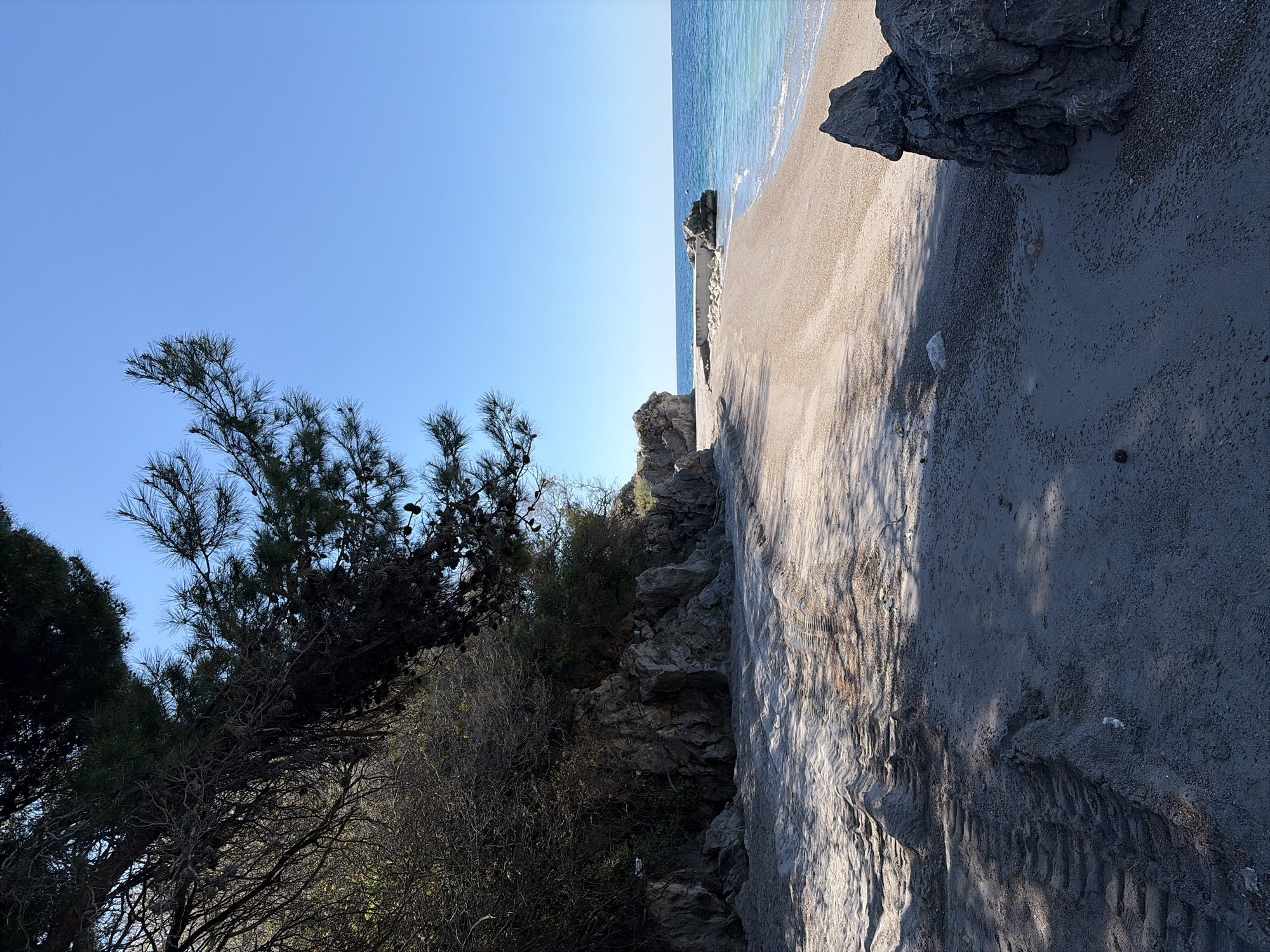 The image depicts a coastal scene in Unknown, Unknown. A beach with light-colored sand is present, meeting a body of water, likely the ocean or sea, with visible waves. Rocky formations are situated along the beach. Vegetation including trees and shrubs grow adjacent to the beach. The sky is blue.
