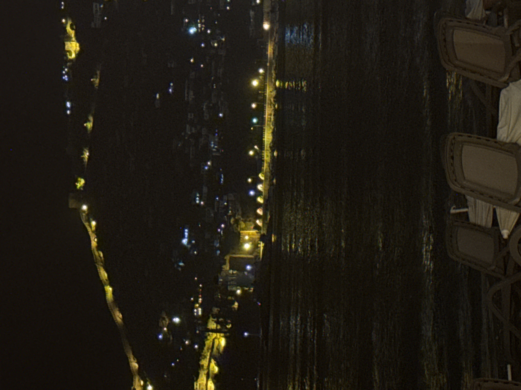 The image depicts a night scene in Alanya, Turkey, focusing on the coastline and the illuminated Alanya Castle on the hill. In the foreground, partially visible are two empty deck chairs and some white fabric, suggesting a seating area overlooking the water. The middle ground shows a body of dark water, likely the Mediterranean Sea, with reflections of lights from the shore. Along the waterfront, a promenade or roadway is visible, lined with streetlights and illuminated buildings, creating a festive atmosphere. In the background, the imposing Alanya Castle, a prominent landmark, is perched atop a steep hill. The castle walls and some structures within it are lit up, casting a warm glow against the dark night sky. The overall impression is one of a tranquil evening in a picturesque coastal city.