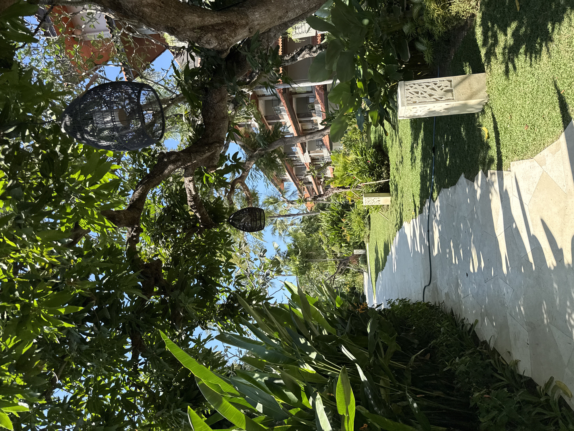 This image captures a sun-drenched pathway in Kuta Selatan, Indonesia, leading through a lush, tropical resort landscape. On the left, a dense wall of vibrant green foliage, featuring large, broad leaves, lines the path. The pathway itself is paved with light-colored hexagonal stone tiles that cast dappled shadows from the overhanging trees.
To the right of the path, a meticulously manicured lawn stretches out, dotted with square, decorative light fixtures featuring carved patterns. In the background, a multi-story hotel building with terracotta-tiled roofs and balconies is visible, nestled amongst palm trees and other tropical vegetation. Two dark, woven pendant lights hang from the branches of a large tree in the foreground, adding a touch of ambient lighting to the outdoor setting. The clear blue sky suggests a bright, warm day. No people are visible in the scene.