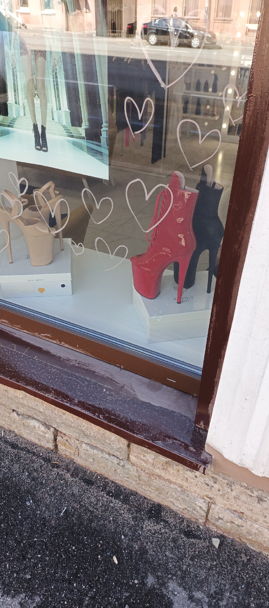 This is a close-up, eye-level, outdoor, full shot of a storefront window display in Saint Petersburg, Russia. The window is reflective and shows a street scene with a black car driving by, as well as some buildings in the background. Inside the window display, there are several pairs of high-heeled shoes. A pair of light tan platform heels are on the left, displayed on a white box. To the right of these, there is a red patent leather stiletto boot with laces and a black stiletto boot. Both of these are also on white boxes. The window itself has several white heart-shaped decals on it. The window frame is dark brown, and the sill below is made of rough, tan-colored stone blocks. The ground in front of the window is dark asphalt with scattered small pebbles.