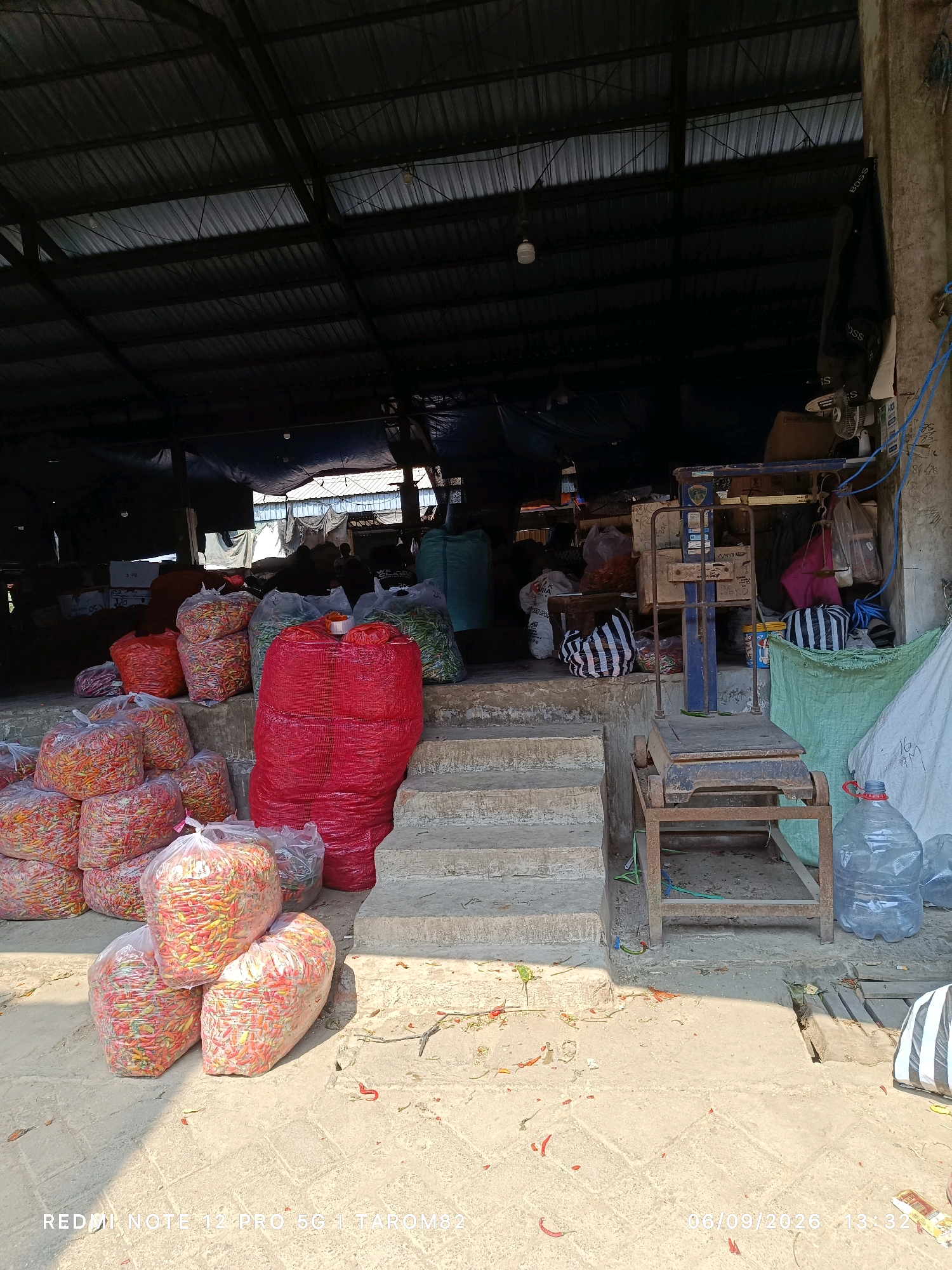 The image depicts an outdoor market stall, likely in Pare, Indonesia, filled with produce, predominantly chili peppers. Several large, red sacks and clear plastic bags are piled high with vibrantly colored chili peppers, varying in shades of red, orange, and yellow. In the background, a covered structure with a corrugated metal roof provides shelter, supported by concrete pillars. A rusty weighing scale stands near the entrance of the stall, suggesting a place of commerce. The ground is paved with concrete tiles, littered with a few scattered chili peppers and dried leaves. The scene is illuminated by natural daylight, casting distinct shadows. The overall impression is that of a busy agricultural market, ready for trade.