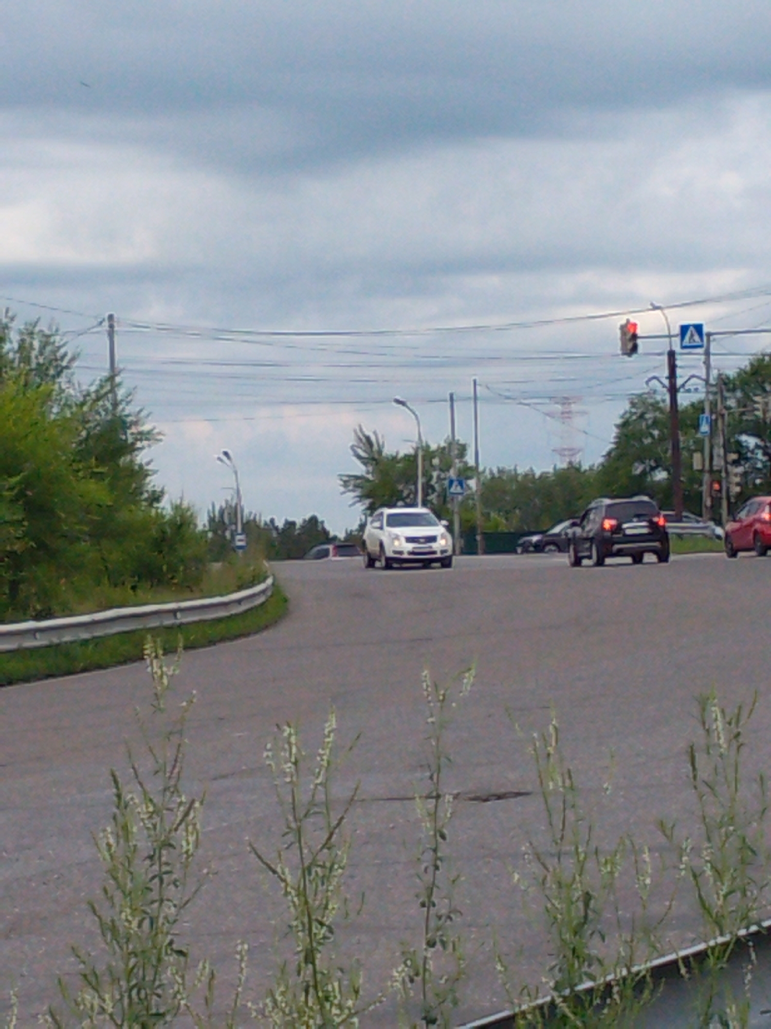 The image shows a traffic intersection with several cars. A white sedan is in the foreground, followed by a black SUV. A red car is visible on the right, and another white car is in the distance. There are traffic lights and road signs present, indicating it is a controlled intersection. Trees and foliage line the sides of the road. The sky is overcast. The scene is consistent with a street in the city of Khabarovsk, Russia.