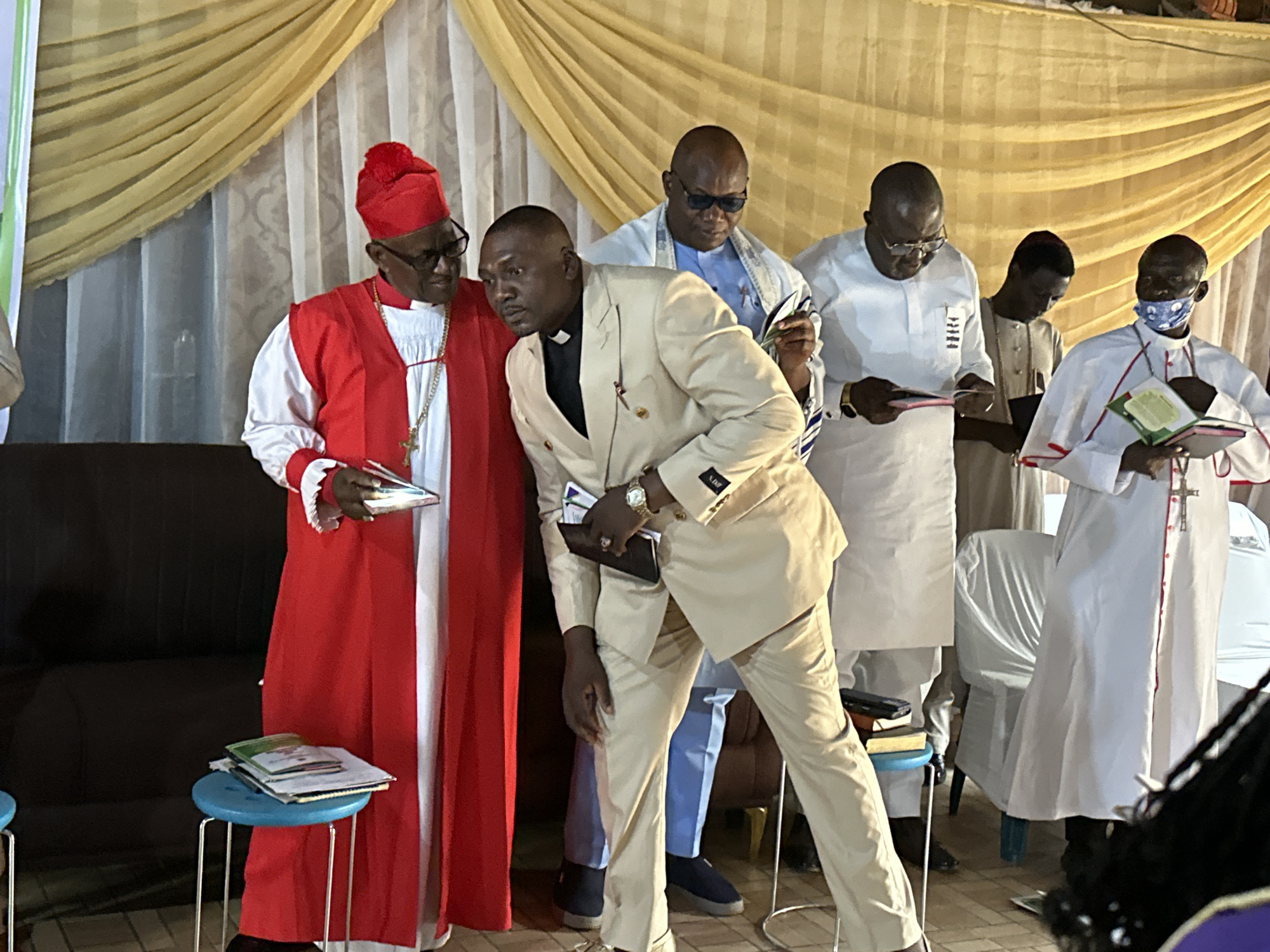 In Barden Yana, Nigeria, at an indoor event featuring a gathering of men in religious attire, a prominent figure on the left wears a vibrant red robe with a white collar and a matching red cap, adorned with a gold chain and cross necklace. He holds a book. To his right, a man in a cream-colored suit leans forward, also holding a book. Behind them, several other men are visible, some in white attire and one wearing sunglasses. In the background, a yellow drapery hangs, and chairs are arranged. On the far right, a person in a white robe with red trim holds an open book, wearing a blue and white patterned face mask. Two blue stools with books are placed near the front left. The lighting suggests an indoor setting.