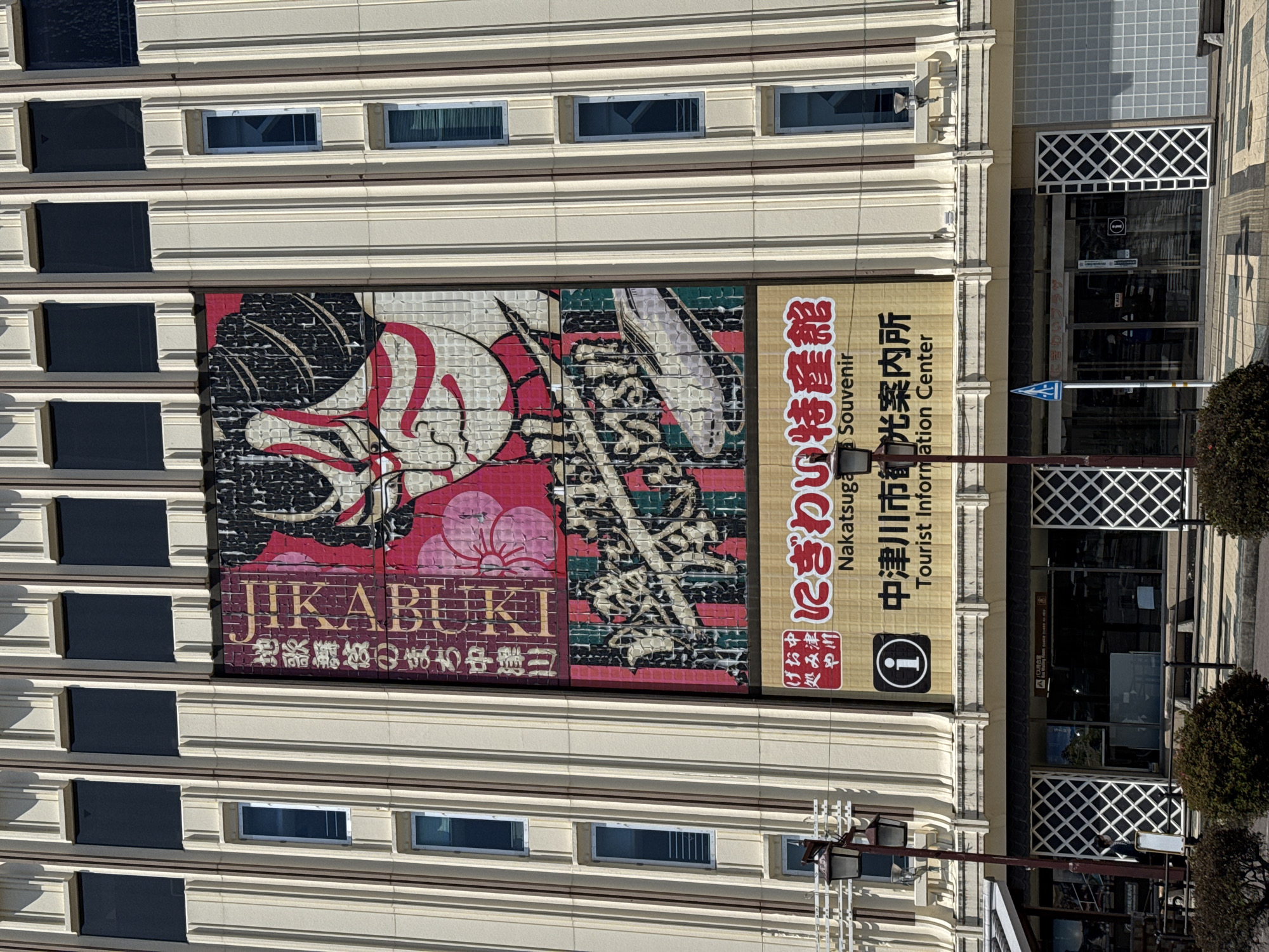 The image shows a multi-story building in Nakatsugawa, Japan, featuring a large advertisement for "Jikabuki", referring to local traditional kabuki theater. The advertisement depicts a kabuki mask and Japanese script. Adjacent to the kabuki ad is signage for the "Nakatugawa Souvenir Tourist Information Center". The building's architecture includes a facade with rows of windows and a decorative lattice design around the entrance. A street sign is visible near the entrance. The building appears to be located on a street with trees.