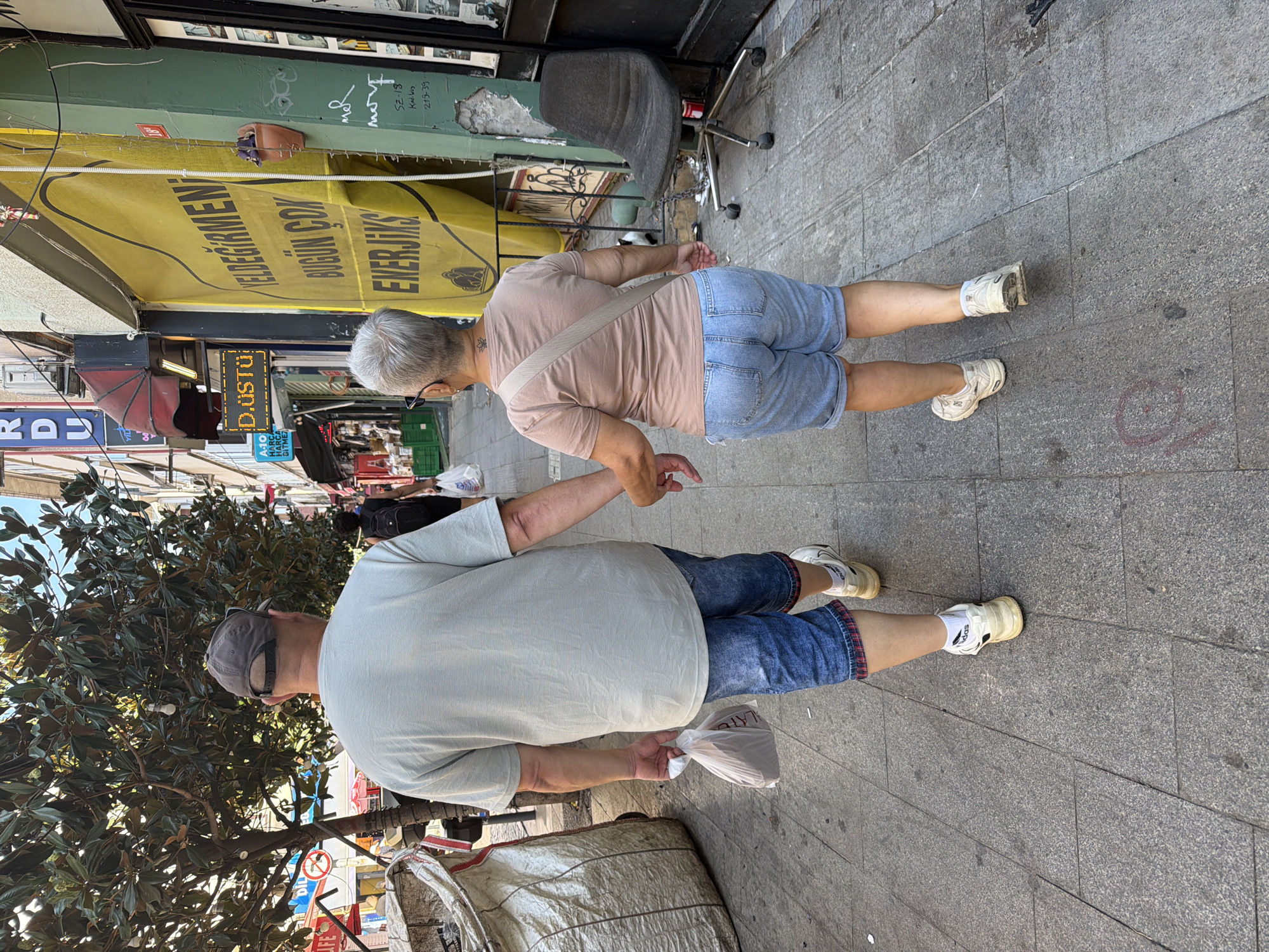 An elderly couple walks hand-in-hand down a sidewalk in Kadıköy, Turkey. The man, on the left, wears a grey t-shirt, blue denim shorts, and a grey baseball cap. He carries a white plastic bag in his left hand. The woman, on the right, wears a peach-colored t-shirt, light blue denim shorts, and a beige crossbody bag. Both wear white sneakers. They are walking away from the camera.
The sidewalk is paved with grey granite tiles. To the left of the couple is a large, weathered, light-colored sack or tarp. Behind them, various shops line the street. A bright yellow banner with Turkish text is prominent on a building to the right, partially obscuring a green wall and a black office chair on wheels. The scene appears to be a bustling commercial street during daylight hours.