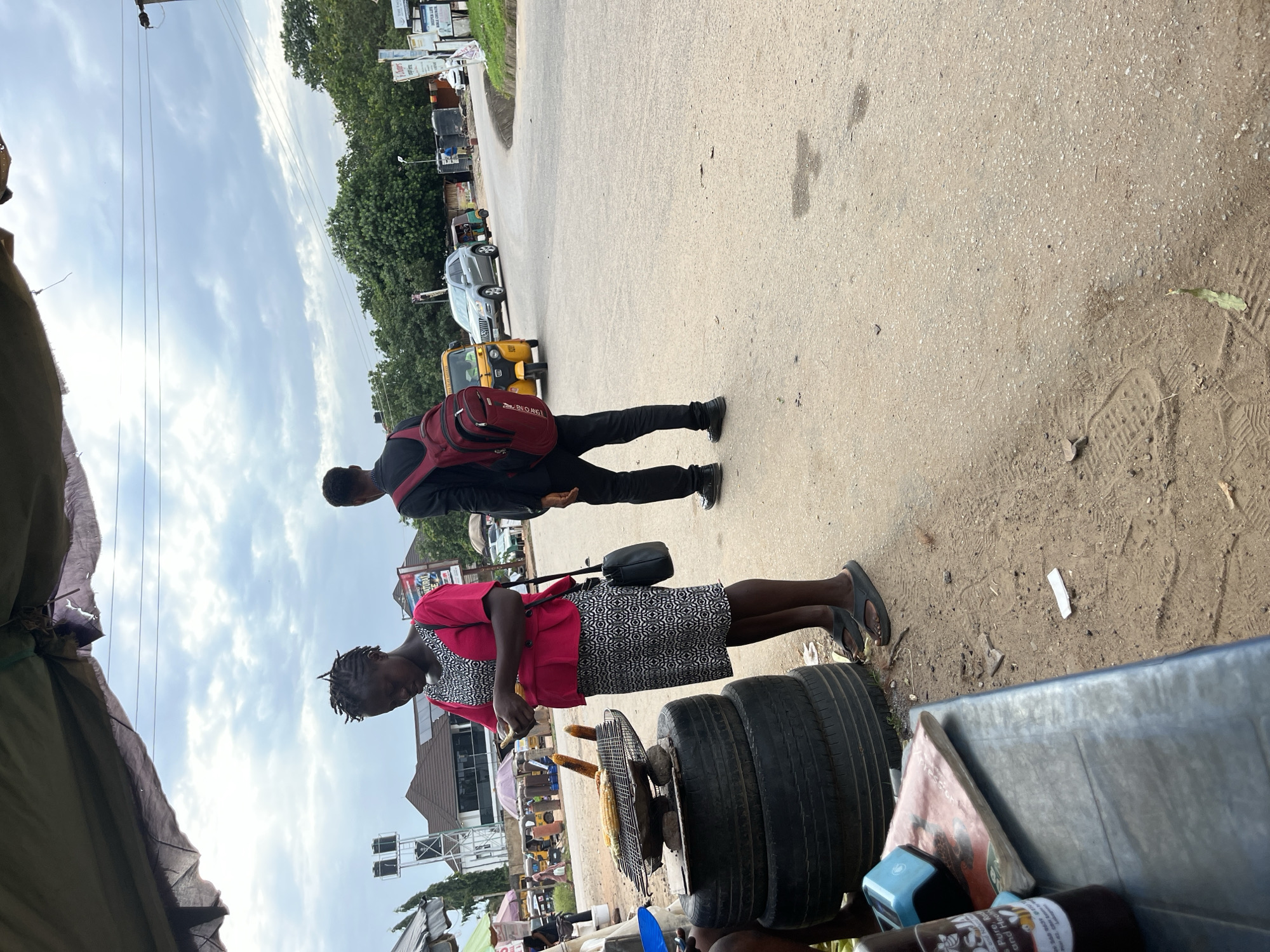 A street scene in Abuja, Nigeria, shows a woman in a pink top and patterned dress standing next to a grill with corn. She is holding a mobile phone. A young man wearing a black suit and a red backpack stands to her right. Several vehicles, including a yellow auto-rickshaw and a silver SUV, are visible on the road. The scene is set against a backdrop of trees and buildings under a cloudy sky.