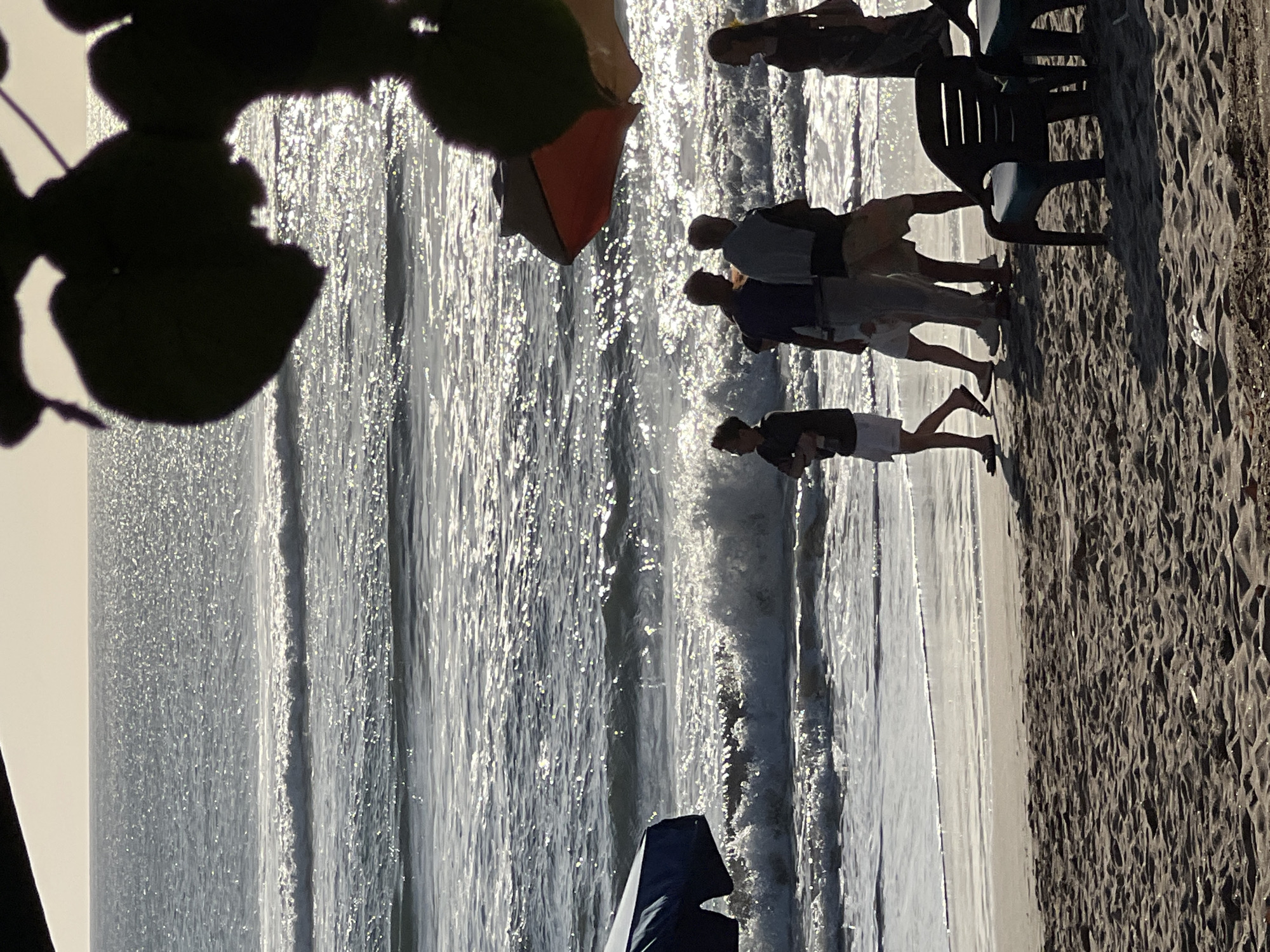 In Kuta, Indonesia, a group of people are walking along the beach as waves crash on the shore. A blue umbrella is visible to the left of the frame, and a red and orange umbrella is angled from the upper right. Dark foliage hangs over the scene from the top. The ocean is shimmering under bright sunlight, with foamy waves rolling in towards the sandy beach. The people, silhouetted against the bright ocean, are casually dressed and appear to be enjoying the beach setting. Two chairs are partially visible in the foreground, casting shadows on the sand.