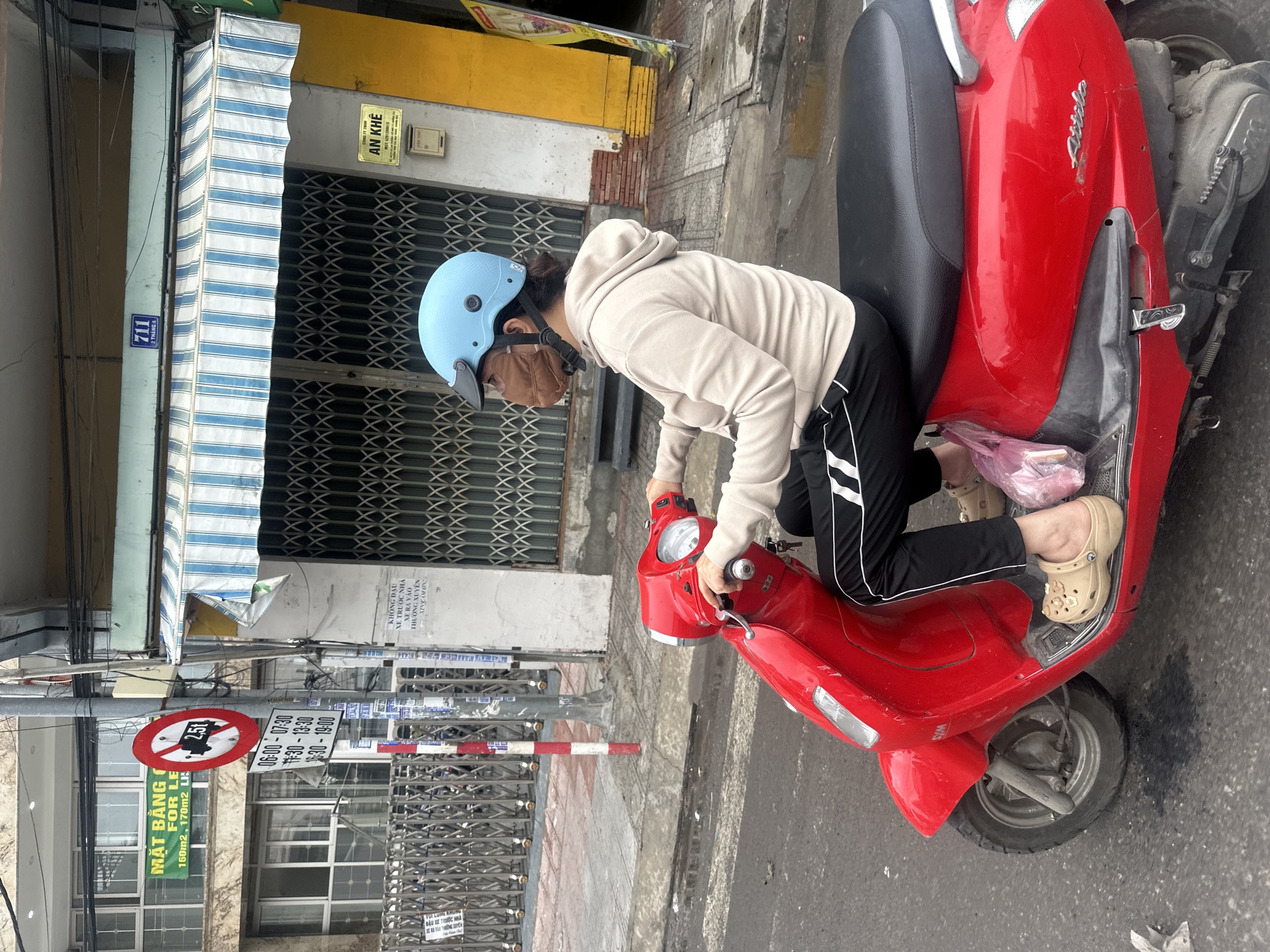 A woman wearing a light blue helmet and brown face mask is seated on a red scooter on a city street. She wears a beige hoodie and black pants with white stripes. The scooter appears to be an older model with some damage. The street scene includes buildings with metal roll-up gates and a striped awning. A street sign with a red circle and a white bar across it is visible, along with a sign with Vietnamese text and time notations (6:00-7:30, 11:30-13:30, 16:30-19:00). Another sign indicates "711 2 THÁNG 4" and "AN KHÊ," suggesting a location in Bac Nha Trang Ward, Vietnam. The ground appears to be paved and slightly wet. The woman is holding the scooter's handlebars as if about to drive.