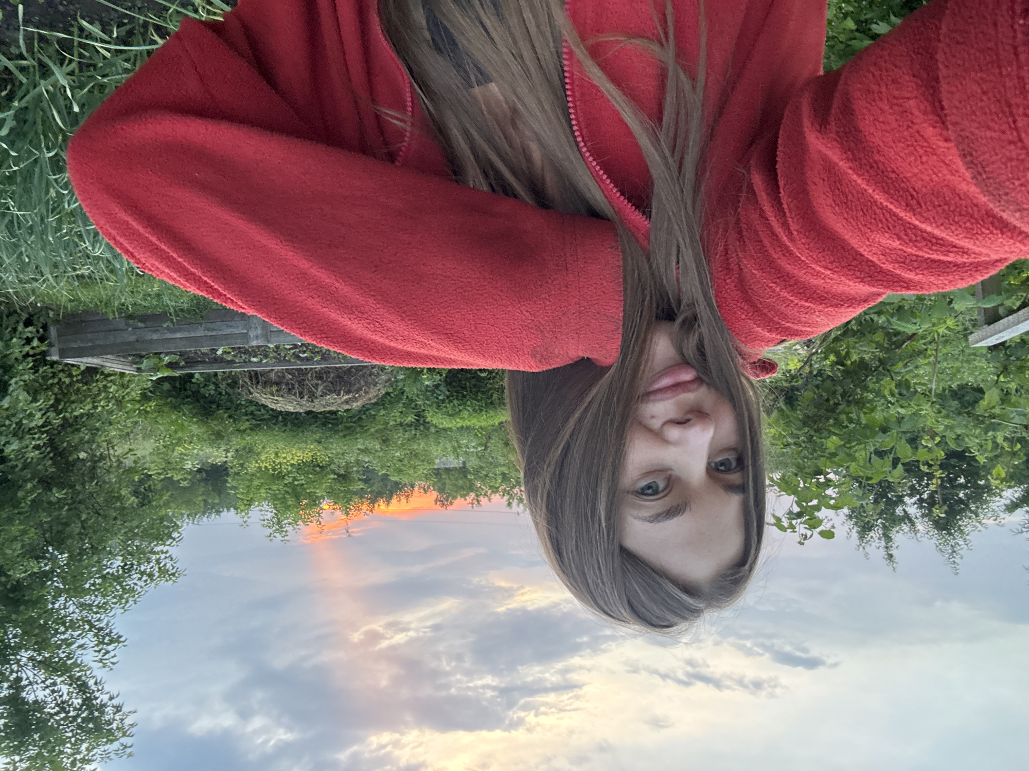 A young woman with long brown hair is taking a selfie in an outdoor setting. She is wearing a red zip-up jacket over a dark garment. Her left arm is bent, and her hand is touching her face. She is looking directly at the camera with a slight smile. The background consists of lush green foliage and trees. In the distance, a strip of orange and pink light is visible in the sky, suggesting either a sunset or sunrise. A raised garden bed and a pile of organic material are also present in the background, indicating a rural or garden environment. The scene appears to be in a natural, unurbanized area.