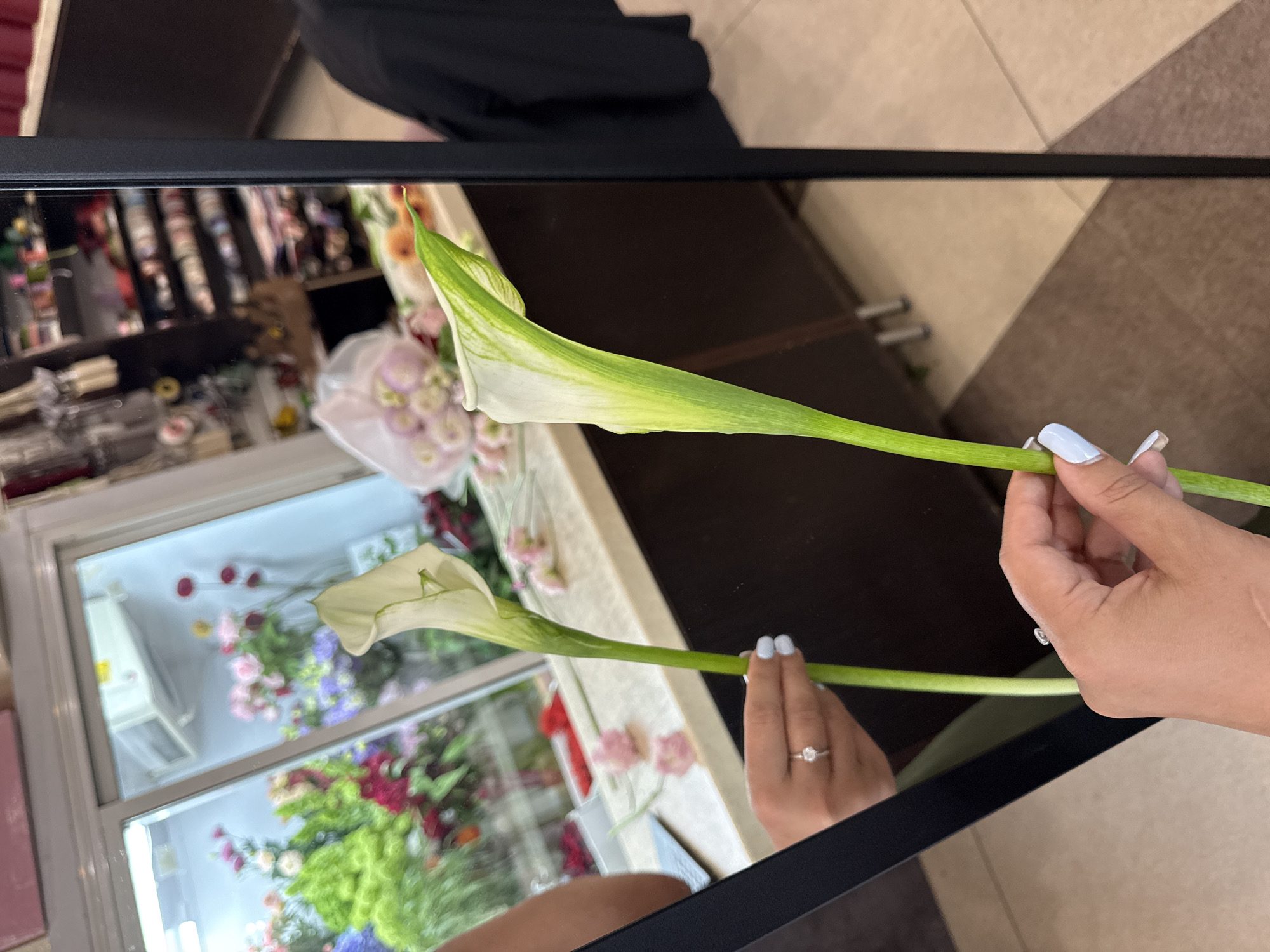 A person holds a calla lily with white and green petals and a long green stem. The person's hand is adorned with a ring and pale blue nail polish. The scene is reflected in a mirror, revealing a flower shop interior with shelves of floral supplies and arrangements of colorful flowers. The floor is tiled with cream and brown squares. The mirror itself is framed by a black border. The image does not provide any information about the city of Assinovskaya, Russia.
