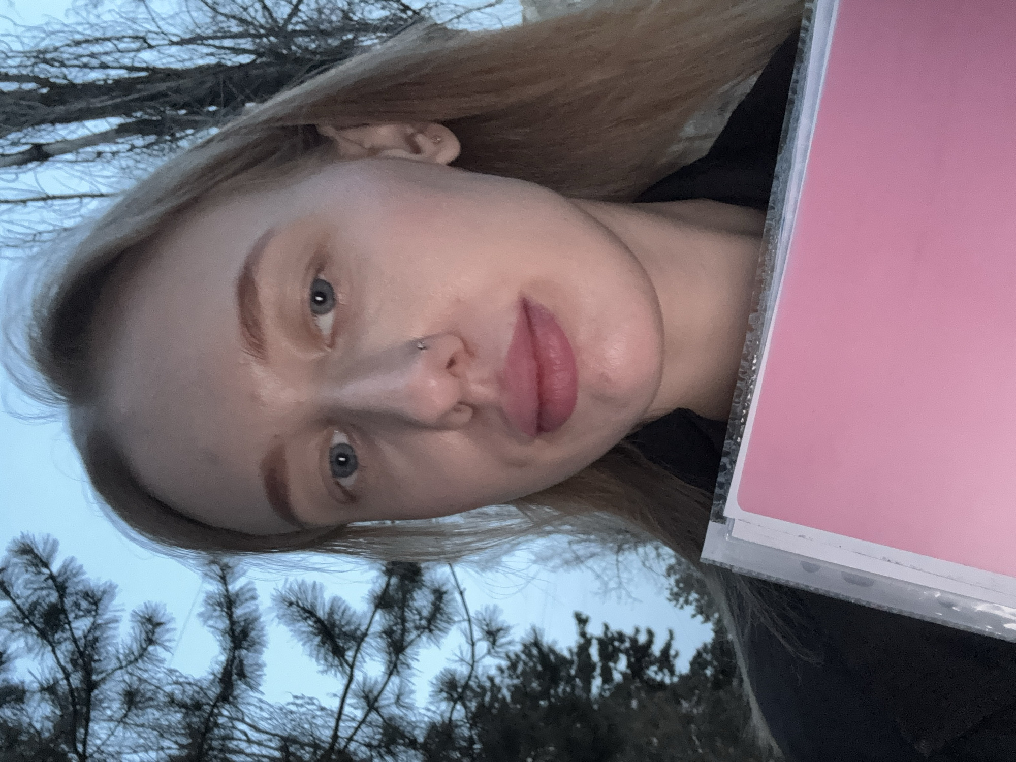 A selfie of a fair-skinned person with blonde hair. The person has blue eyes and a nose piercing. They are holding a pink document or object in a plastic sleeve. The background includes trees and a pale sky, suggesting an outdoor setting. There's no definitive evidence indicating the photo was taken in Azov, Russia, or any specific location.