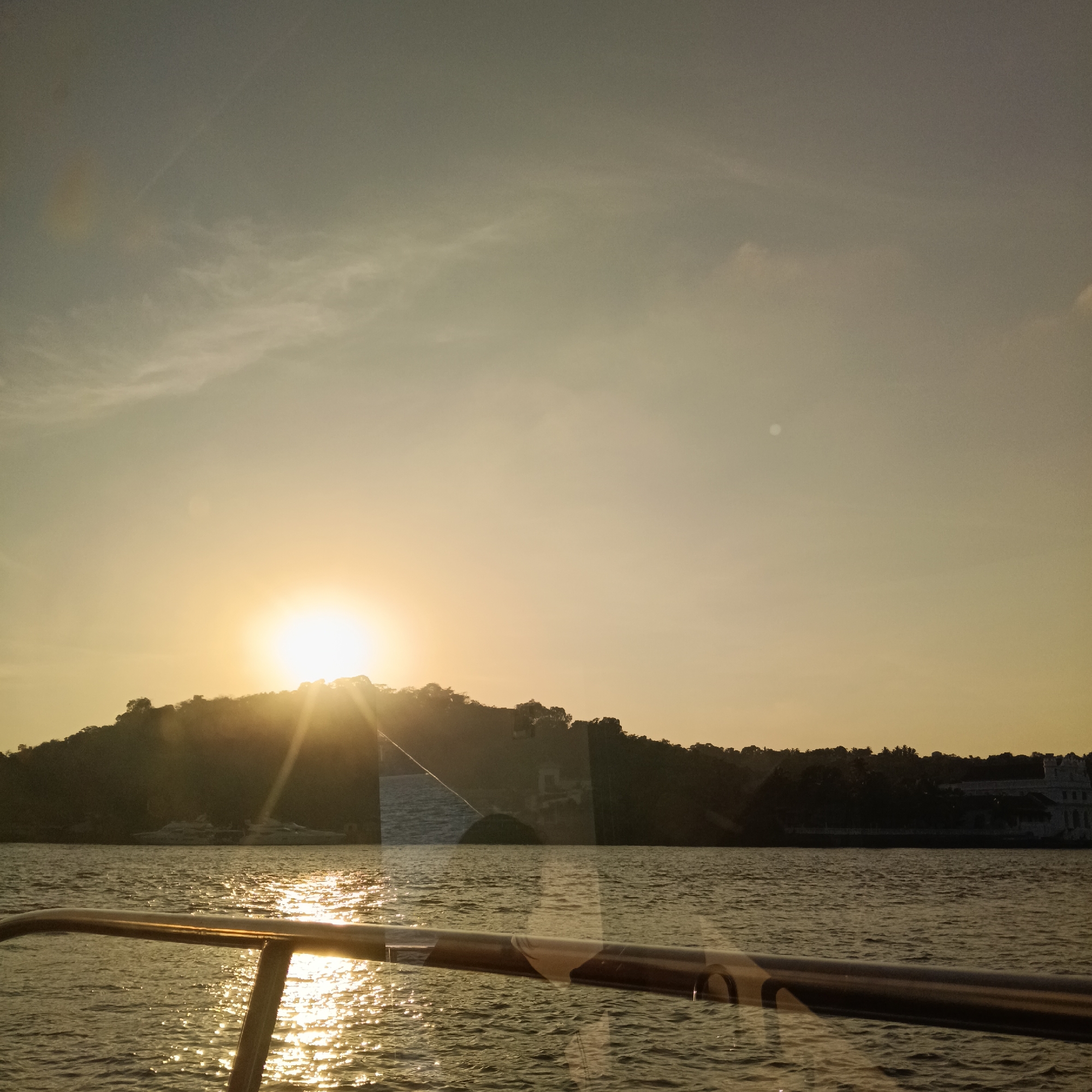 The image captures a sunset over the Mandovi River in Britona, Goa, India, as viewed from a boat. The sun is setting behind a tree-covered hill, casting a golden light across the water and sky. Sunlight reflects off the river's surface. A portion of the boat's railing is visible in the foreground. On the opposite bank, there is a building with white walls. A couple of boats are docked on the bank across from the photographer.