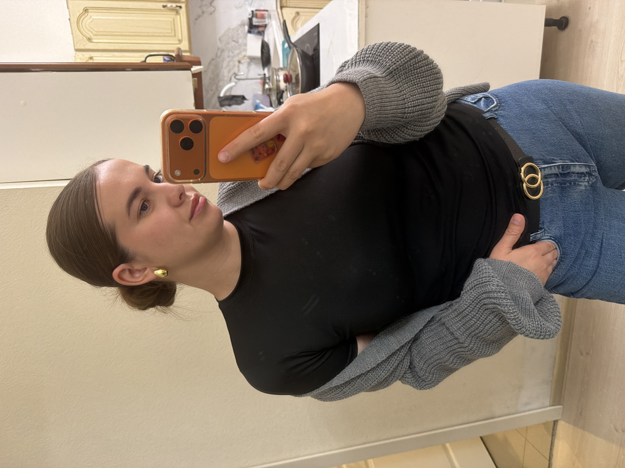 A young woman is depicted taking a selfie or a mirror self-portrait in an indoor setting in Sochi, Russia. She holds an orange-cased smartphone, featuring three visible camera lenses on its back, in her right hand. The phone is angled towards her face, capturing her reflection or a direct shot.
The woman has light skin, dark hair pulled back into a neat bun, and wears a neutral, relaxed expression as she looks towards the phone. She is dressed in a black fitted long-sleeved top, light blue jeans, and a grey knitted cardigan. A distinctive black belt with two interlocking gold-colored rings as a buckle is prominent around her waist. A small, gold-colored hoop earring adorns her left ear.
The background indicates a casual home environment, likely a kitchen or an adjacent room. Elements such as light-colored cabinetry, some with decorative detailing, and what appears to be a kitchen counter or appliance are visible. The lighting is artificial, consistent with an indoor setting. There is no visible text in the image.