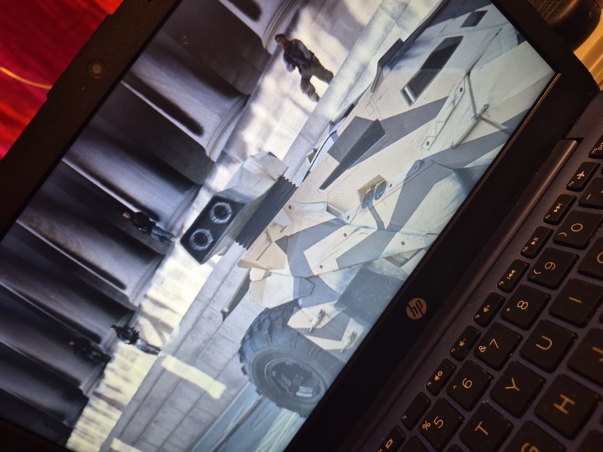 A laptop displays a scene of a futuristic military vehicle with a camouflage paint scheme, resembling a heavy armored car, parked on a paved surface. The vehicle has large, rugged tires and what appears to be a double-barreled weapon mounted on top. In the background, several figures, likely soldiers in tactical gear, are visible standing near large architectural pillars, suggesting an urban or historical setting. The overall image is a close-up of the laptop screen, with the keyboard visible in the foreground, suggesting the laptop is open and in use. The scene on the screen appears to be from a video game or a movie. There is no information within the image to suggest any connection to Sandwell, United Kingdom.