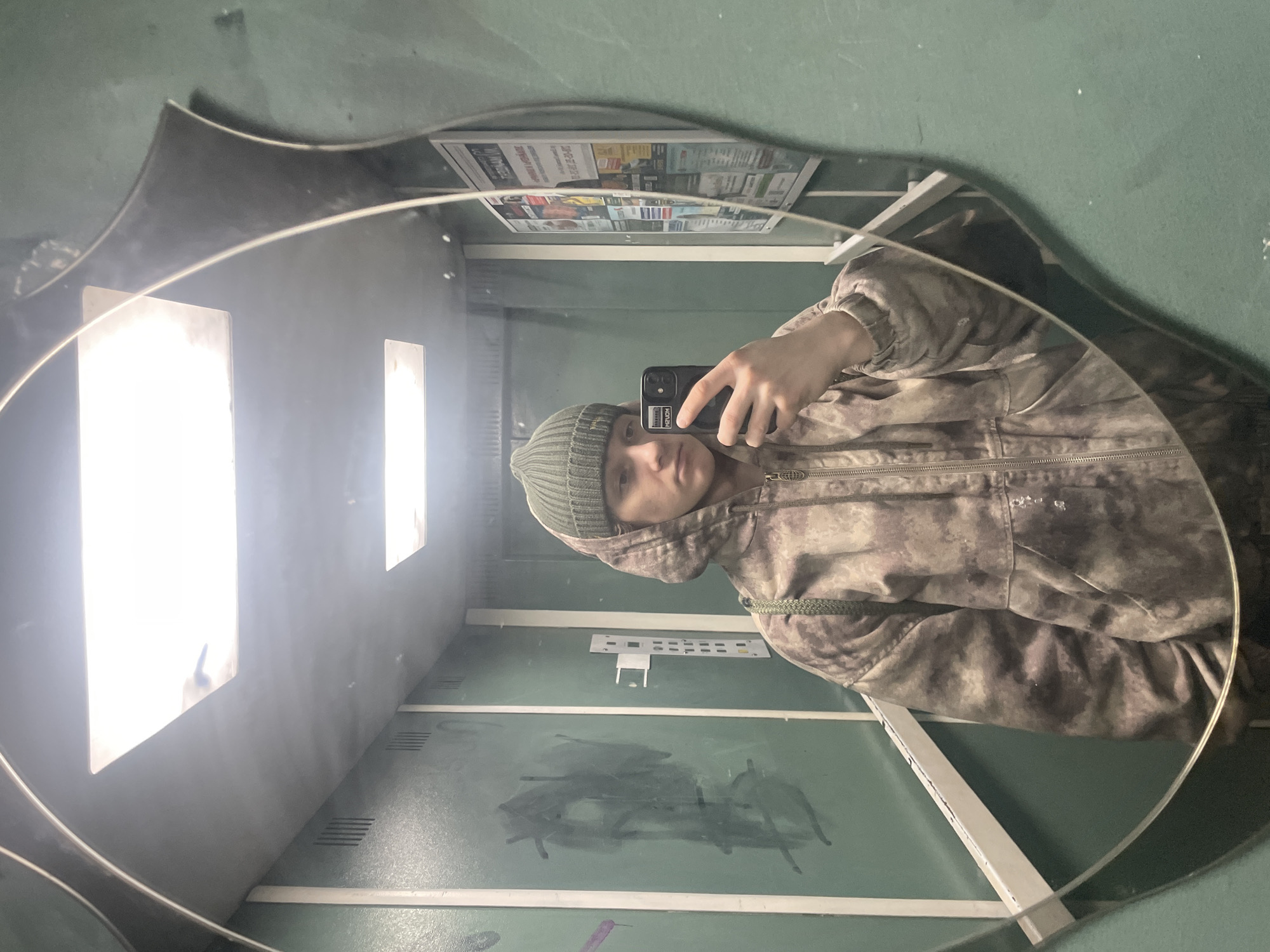 A person wearing a camouflage jacket and a green beanie is taking a selfie in a mirror with an irregular oval shape. The reflection shows the interior of what appears to be a building, potentially a public restroom or industrial facility, with pale green walls and a gray floor. Graffiti-like markings are visible on the wall in the reflection. Above the person's head in the mirror, there is a poster of some sort. The location is identified as Krasnoyarsk, Russia.