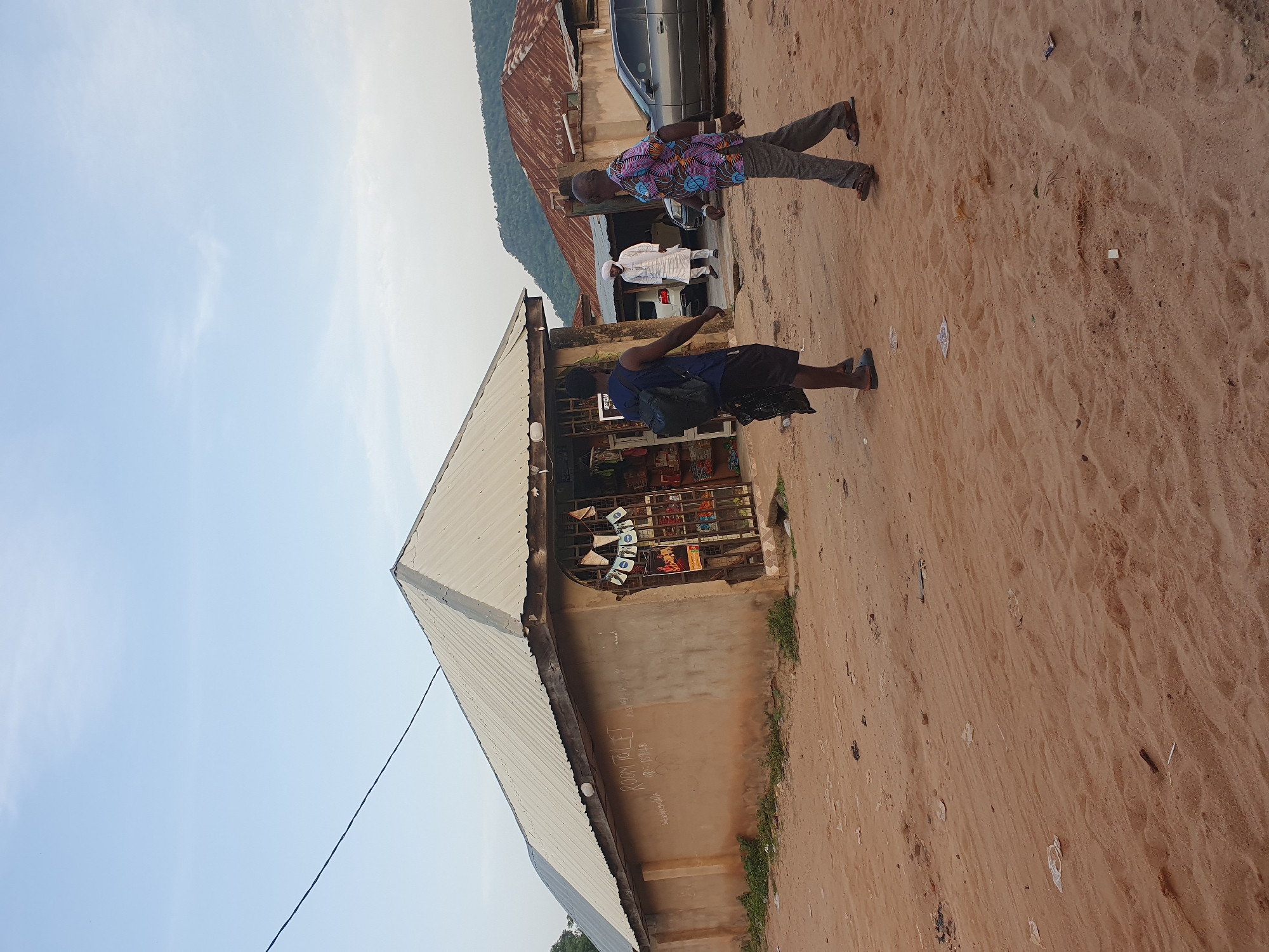 In Lokoja, Nigeria, an outdoor scene depicts a dirt path in front of a small, stucco-walled shop with a corrugated metal roof. Two men are walking on the path in front of the shop. The man on the left, wearing a blue shirt, backpack, and dark shorts, walks away from the camera. The man on the right, wearing a colorful patterned shirt and grey pants, walks toward the camera. Behind the shop, a man in white clothing stands near a parked white car. Further in the background, a grey car is parked next to a building with a rusty corrugated metal roof. A verdant hillside is visible in the far distance. The ground is covered in sand and scattered debris.