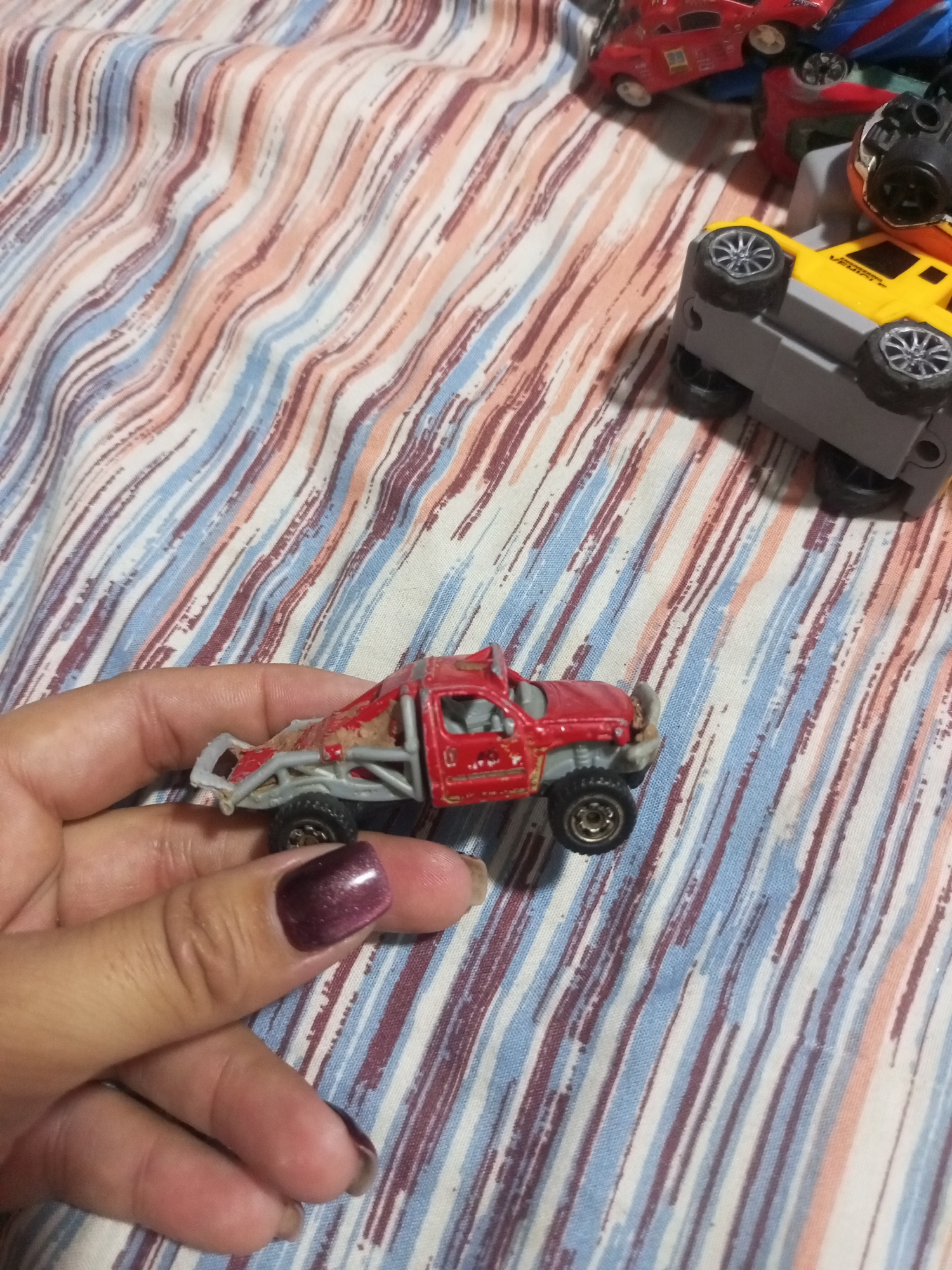 A close-up, eye-level, outdoor shot focuses on a person's hand holding a small, red, toy off-road vehicle. The vehicle appears to be made of plastic and metal, with a roll cage and large tires. The hand has painted fingernails with a metallic burgundy polish. In the background, a striped fabric, possibly a blanket or bedsheet, features an abstract pattern of wavy, vertical lines in shades of white, light blue, pink, and dark red. Two other toy vehicles are partially visible: a yellow and gray truck overturned on its side, and a red car further in the background. The overall impression is of a casual, indoor setting. The location in La Tagua, Venezuela is not discernible from the image.