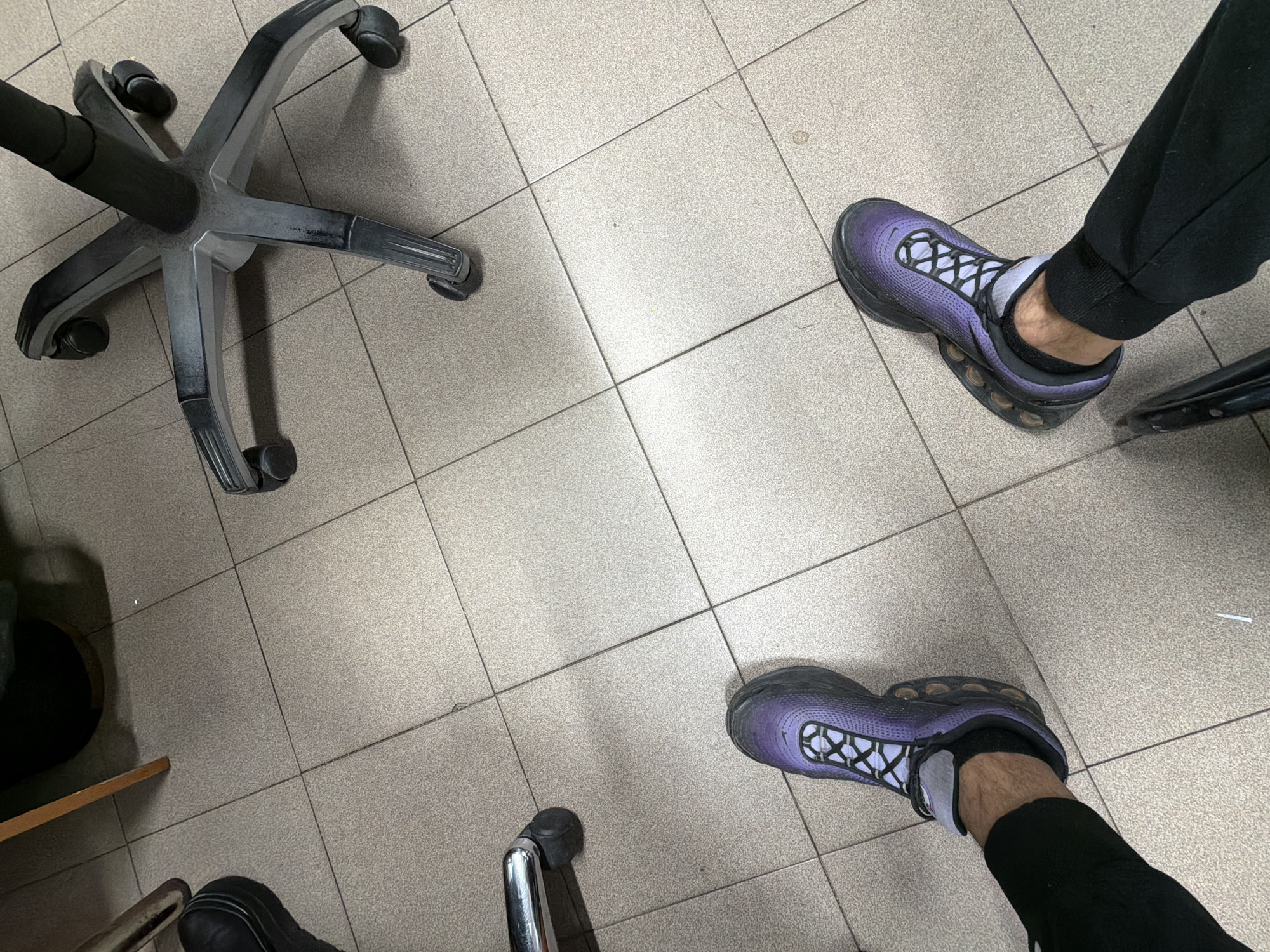 The scene is an indoor setting in Plovdiv, Bulgaria, on a tiled floor. A person is visible with their feet and lower legs, wearing black sweatpants and purple and black sneakers with white socks. An office chair is also visible, with its five-legged base resting on the tile. The floor appears to be a public area.