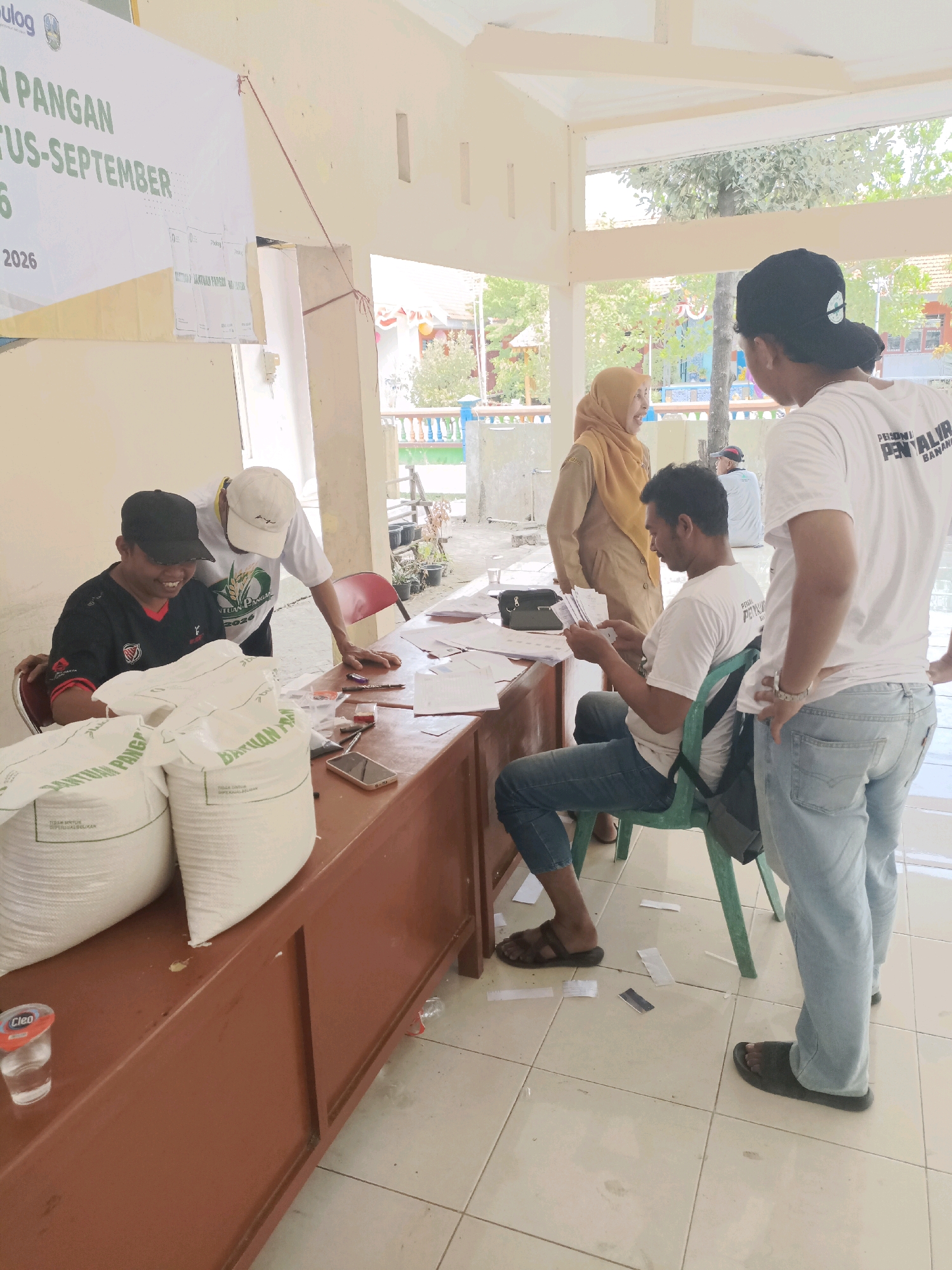In Guminingrejo, Indonesia, a scene unfolds at what appears to be a distribution point for food aid. Two large white sacks, labeled "BANTUAN PANGAN" (Food Aid), sit prominently on a long, brown wooden counter. Behind the counter, a man wearing a black t-shirt and a black baseball cap is seated, smiling and interacting with someone out of frame. Another man, wearing a white t-shirt and a white cap, leans over the counter, looking at papers. To the right, a third man, wearing a white t-shirt with text that appears to read "PERSONIL PENAL," stands and reviews documents he holds in his hands. A woman in a tan hijab and matching long dress stands further back, observing the activity. The environment suggests an outdoor or semi-outdoor covered area, with tiled flooring visible. A banner with Indonesian text, partially obscured, hangs on the wall behind the counter. The overall atmosphere is one of organized distribution and interaction.