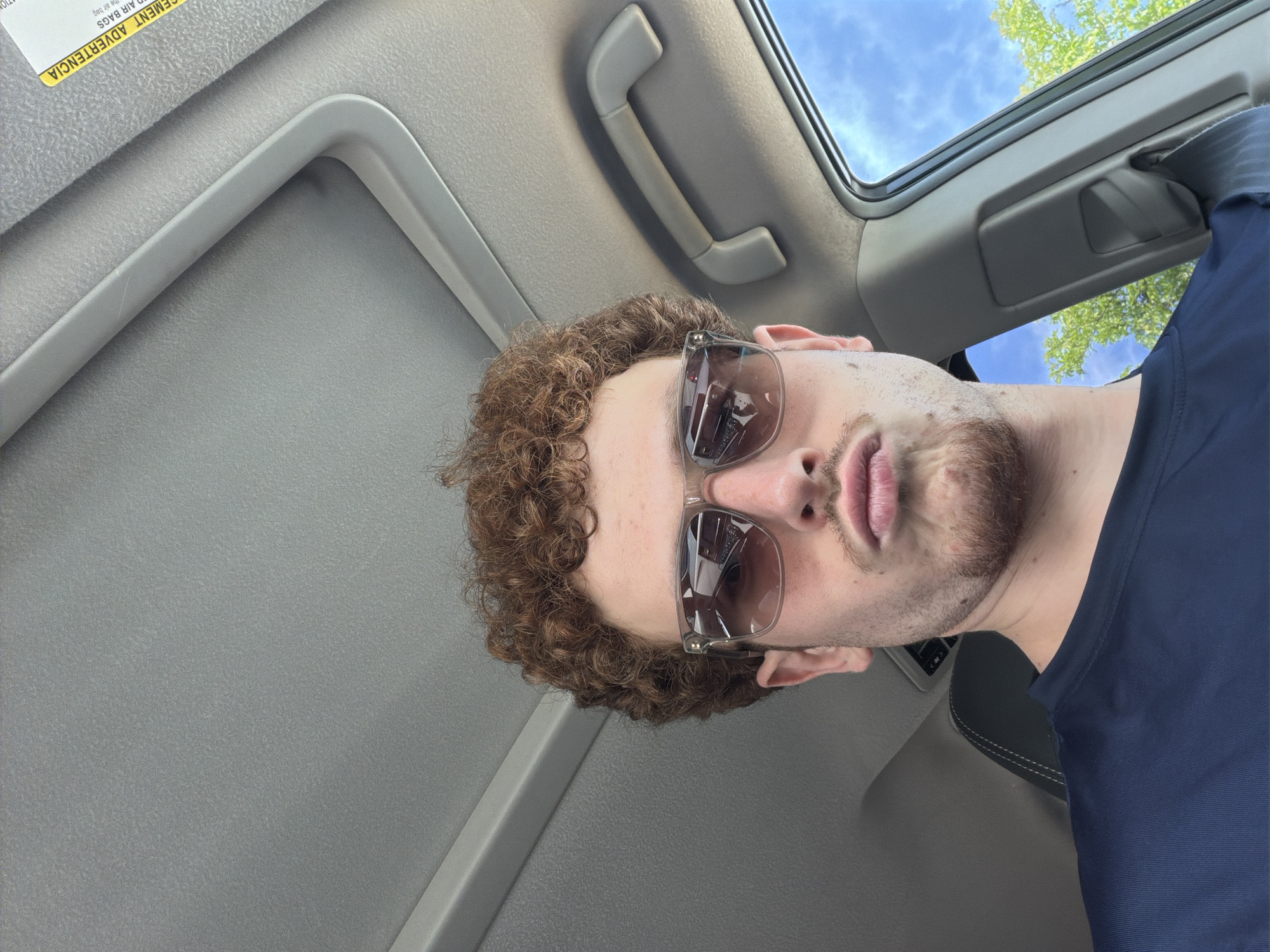A young man with curly brown hair and sunglasses is in the passenger seat of a car. He is wearing a dark blue shirt and has his lips pursed. The car's sunroof is open, revealing a blue sky and green trees. A yellow sticker with text in Spanish is visible on the car's interior. The image was taken in the city of Orange, United States of America.