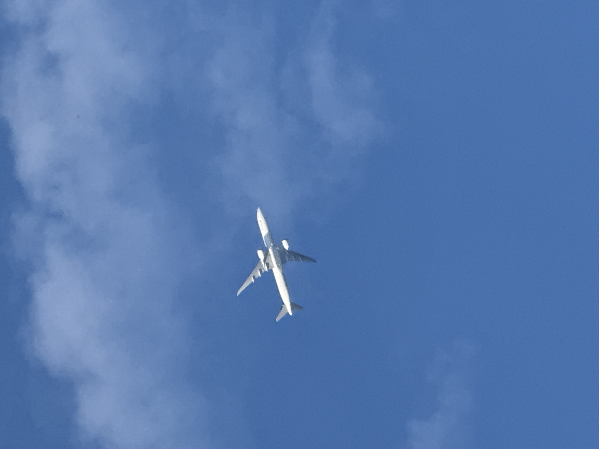 A white airplane is flying against a backdrop of a blue sky with some scattered white clouds. The airplane is viewed from below, showing its undercarriage, wings, and tail. No specific information about the location in London or any people or interactions is available in the image.