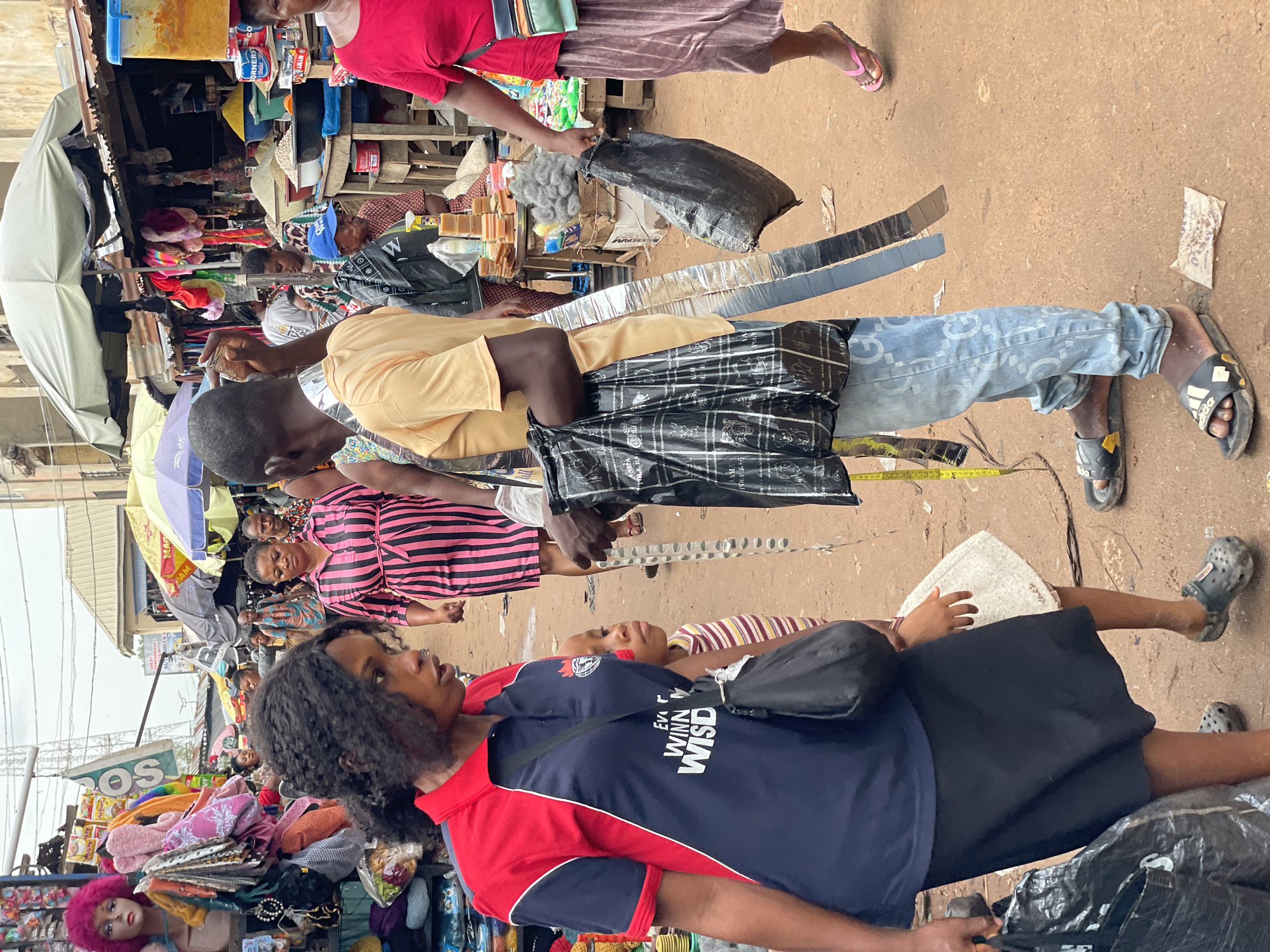 This image captures a bustling street scene, likely a market, in Onitsha, Nigeria. In the foreground, a woman with long, dark curly hair wears a two-toned shirt (red and navy) and a black skirt, and carries a black shoulder bag and a black plastic bag. Behind her, a child wearing a horizontally striped shirt is lying down. A man in a tan shirt and patterned light blue jeans stands behind them, carrying shiny metal strips, a measuring tape, and a large black plastic bag. In the background, numerous people navigate the crowded market, and open stalls with various goods are visible. An umbrella provides shade over the stalls. The ground appears to be a mix of dirt and pavement.