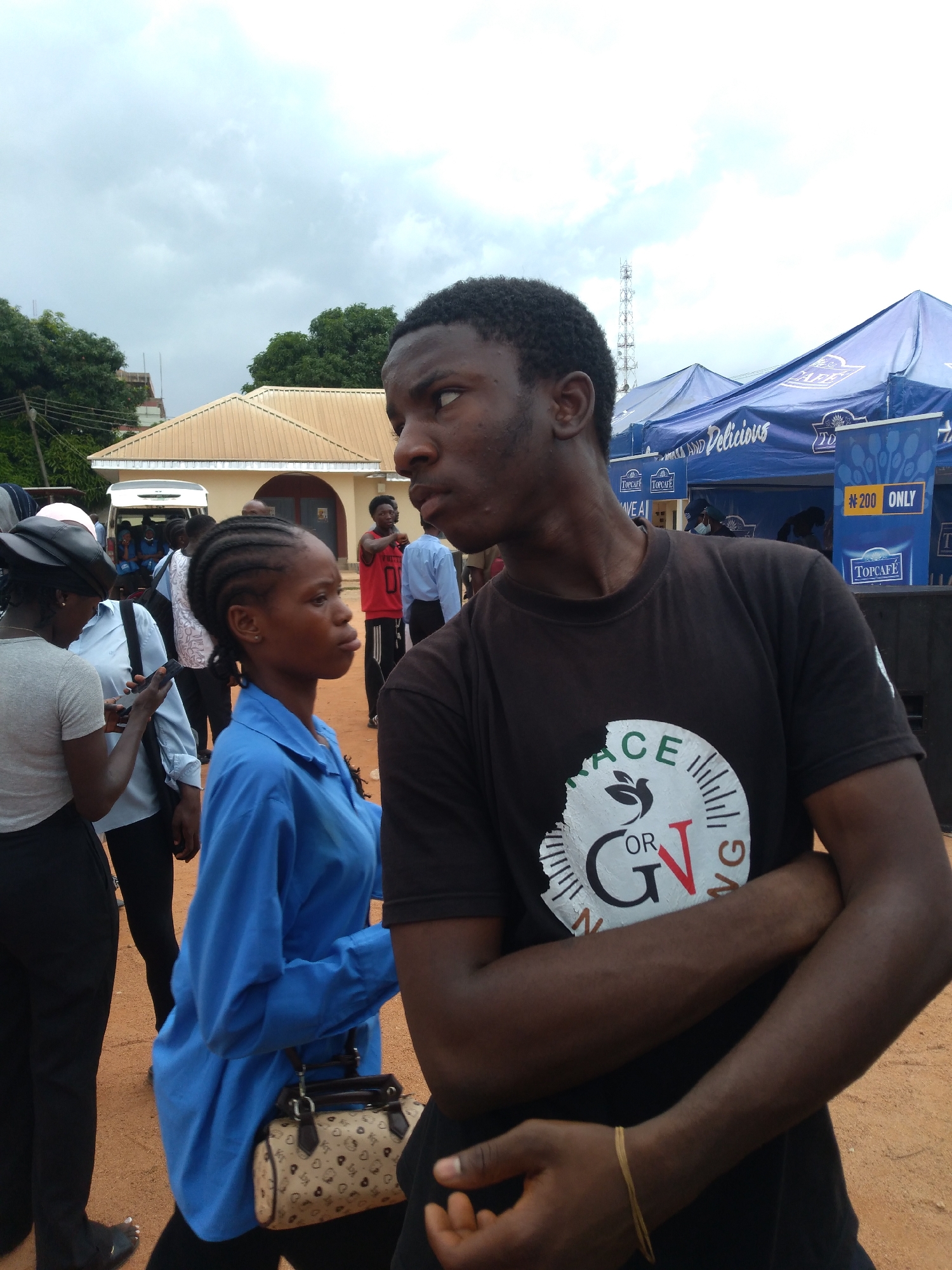 A medium shot captures an outdoor scene on a cloudy day. In the foreground, a young Black man wearing a dark t-shirt with a circular graphic on it has his arms crossed. To his left and slightly behind him, a young Black woman in a blue button-down shirt is also facing to the left, holding a patterned handbag with dark straps. Other individuals are visible in the background, including a person in a red tank top and a person in a light blue shirt. A white van is parked behind some people, and a building with a beige roof and brown trim is situated further back. Blue tents with branding are set up to the right. The ground appears to be dirt. The presence of blue tents with branding suggesting a vendor setup and the general atmosphere imply an event or market setting. The scene is likely in Jos, Nigeria, given the context provided.