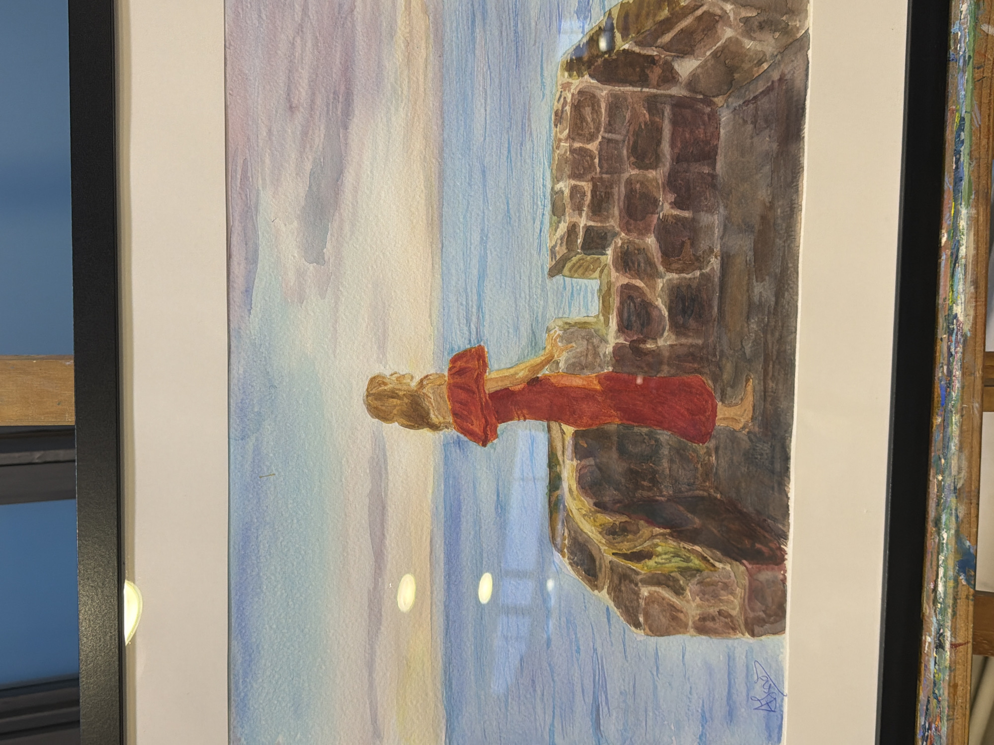 This is a watercolor painting framed in black. The painting depicts a woman in a long, red, off-the-shoulder dress standing on a stone structure overlooking a body of water. The woman is facing away from the viewer, looking out at the horizon. The sky is a gradient of colors, from a light blue at the top to shades of pink and yellow near the horizon, suggesting either sunrise or sunset. The water is a deep blue. The stone structure appears to be old, with rough-hewn stones. There are reflections on the water. A signature, possibly "A. Siso," is visible in the lower left corner of the painting. The painting is displayed on an easel. The background above the painting shows a blue wall and a wooden beam. The context provided indicates the scene is Elche/Elx, Spain, but the painting itself does not contain explicit elements that would identify it as being specifically located in Elche/Elx.