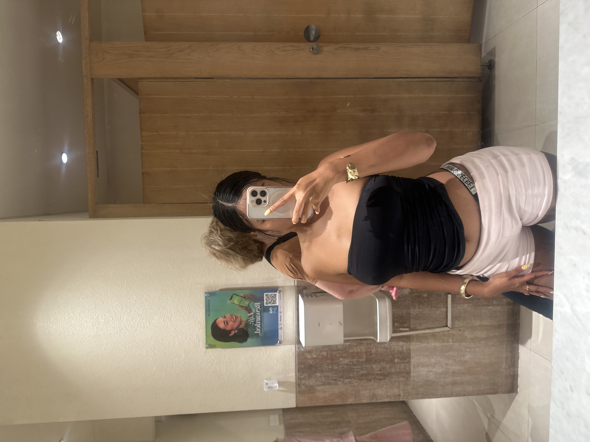 In a restroom with tiled walls and wooden stalls, a young woman is taking a selfie in a mirror. She is wearing a black strapless top and light pink shorts, accessorized with a gold bracelet and rings. A second person, a woman with blonde hair, is partially visible behind her. The restroom contains a hand dryer and a poster on the wall.