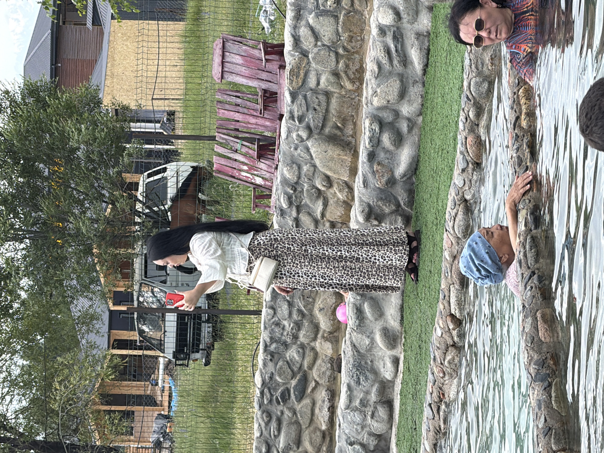 In an outdoor setting with natural stone borders and a water feature, a woman with long black hair and a leopard-print skirt stands and takes a photo with her red smartphone. She wears a white, ruffled blouse and a white crossbody bag. In the background, construction is underway on two wooden structures. A white van is parked behind a metal fence. In the water feature, two other women are visible. One wears a blue headscarf and the other sunglasses and a colorful patterned top.