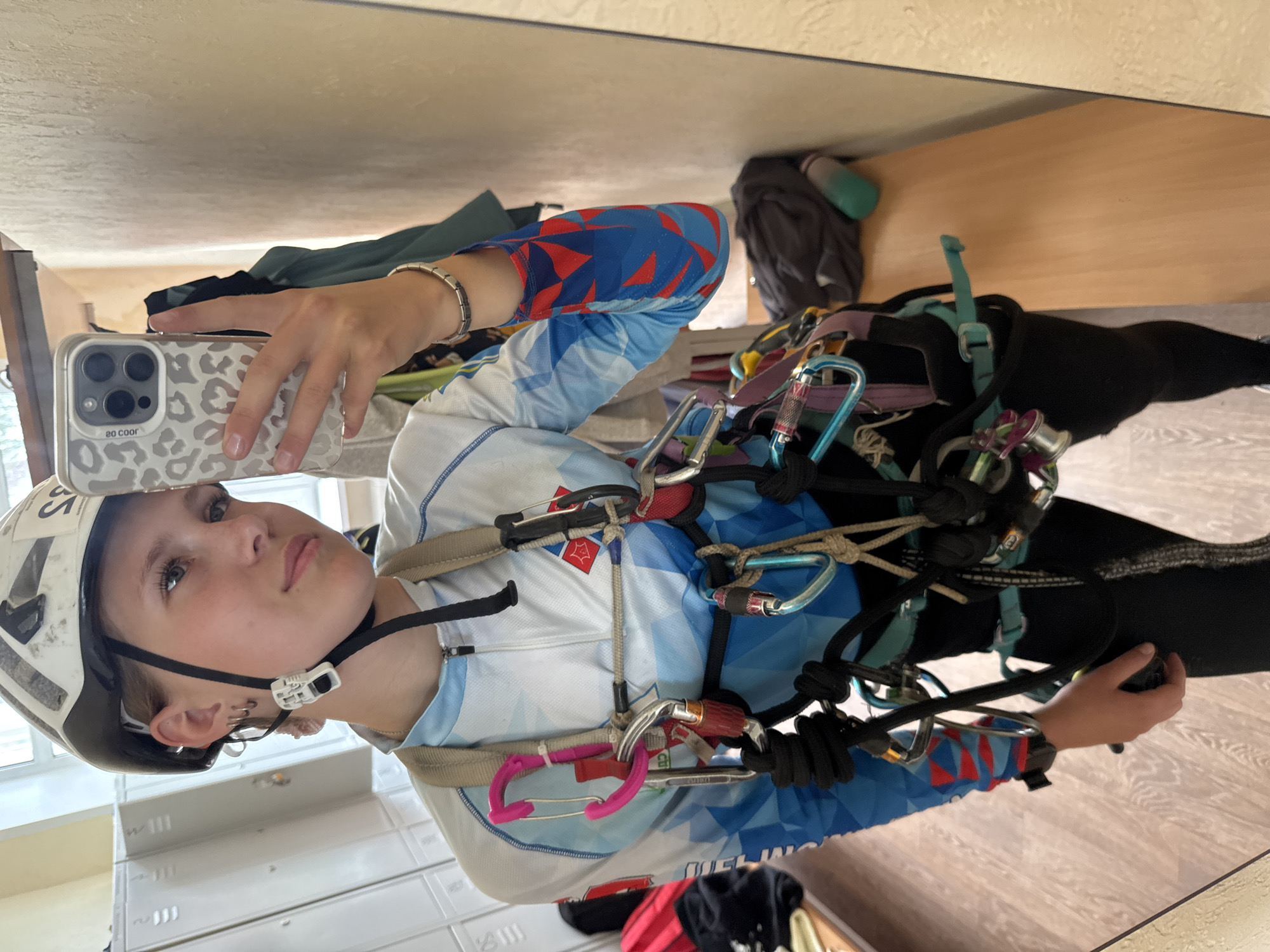 A young person, possibly female, stands in front of a locker and a mirror, taking a selfie with a smartphone. They are wearing a white climbing helmet, a long-sleeved blue and white athletic shirt with a red and blue geometric pattern on the sleeves, and black athletic pants. A climbing harness with various carabiners, ropes, and other gear is worn over the shirt. The environment appears to be a locker room or changing area, with white lockers visible in the background and a wooden bench. There are no other people visible in the image. The location is identified as Chaikovsky, Russia.