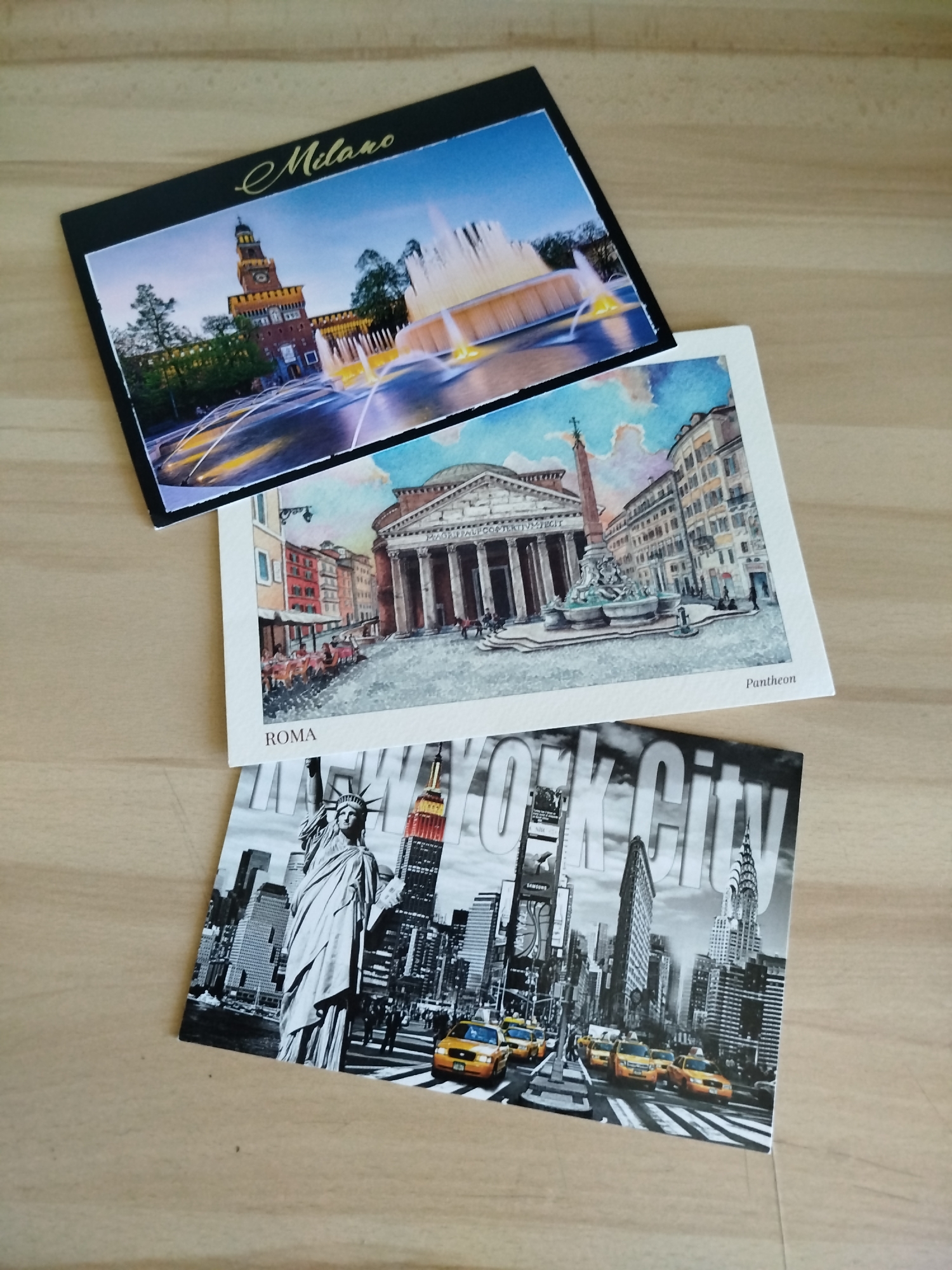 Three postcards are on a light brown wooden surface. The top postcard shows a night scene in Milan, Italy with a castle and illuminated fountains. The middle postcard is a watercolor painting of the Pantheon in Rome, Italy. The bottom postcard is a black and white image of New York City, featuring the Statue of Liberty and yellow taxis. The locations depicted are Milan, Italy; Rome, Italy; and New York City, USA. The image does not contain the city of Varna, Bulgaria.