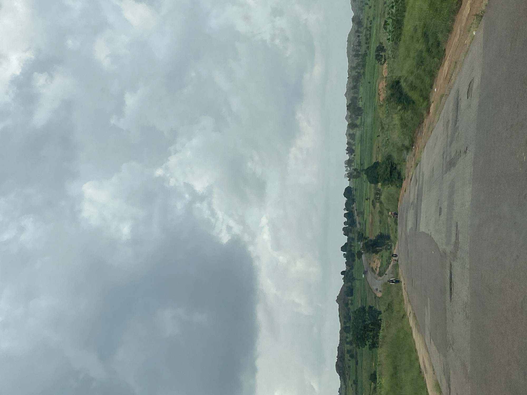 An eye-level, medium shot captures a paved road in Kuru, Nigeria, curving into the distance. The sky is overcast with dense, gray clouds. The road surface is aged, showing patches of wear and tear. On the left side of the road, tall, dry grass grows along the shoulder. On the right, the land slopes upwards, covered in vibrant green vegetation and agricultural fields, interspersed with trees. In the mid-distance, a few figures are visible. One person walks on the road, and two motorcycles are also present. Further back, more trees and rolling hills dotted with vegetation complete the landscape. The scene depicts a rural or semi-rural environment under a cloudy sky.