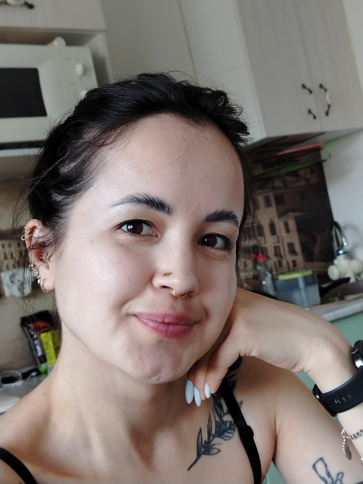 A young adult woman is featured in a close-up, eye-level selfie taken indoors. She has dark hair styled back, dark brown eyes, and a clear complexion, with a subtle, relaxed smile on her face. Her head is tilted slightly to her right, and her left hand gently supports her chin and cheek.
She displays multiple piercings: a prominent gold hoop in her right nostril, a smaller gold stud on her left nostril, and several intricate gold earrings and ear cuffs on her left ear. Tattoos are visible on her left shoulder and upper arm, including a detailed leafy branch on her chest/shoulder. Her fingernails are painted a light, pale blue or white color, and she wears a black smartwatch on her left wrist. A black strap, likely from a top or bra, is visible on her shoulder.
The background suggests a kitchen environment. On the left, a white microwave oven sits on a counter. Above her right shoulder, white kitchen cabinets with dark handles are visible. Behind her, part of a wall features what appears to be a framed picture depicting an old building, possibly a European cityscape. A kitchen sink and faucet are also partially seen in the background. The lighting is bright, indicative of daytime. The woman appears relaxed and content. There is no legible text visible in the image.