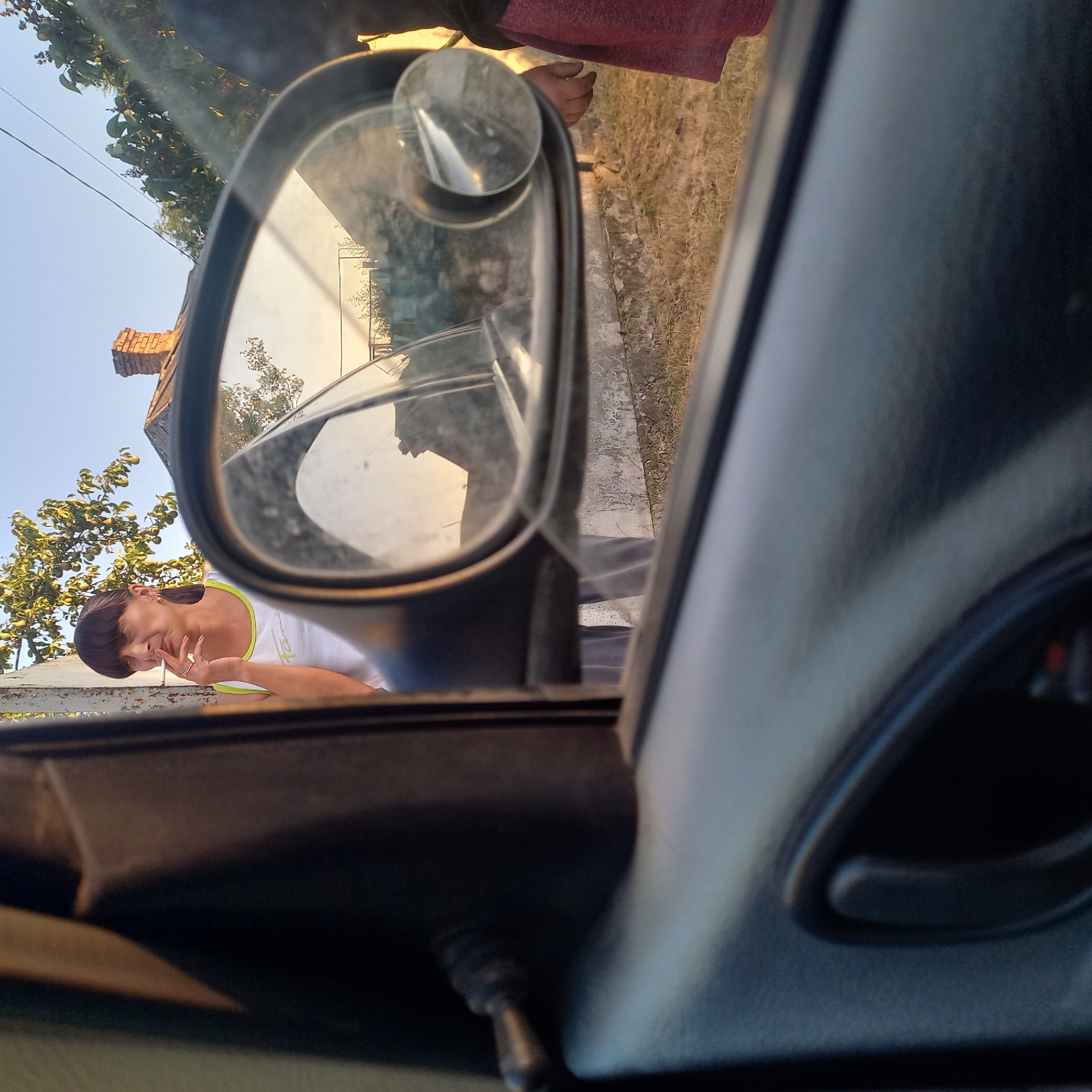 A woman is standing outside a car, smoking a cigarette. She is wearing a white tank top with a green trim and has her hair pulled back in a ponytail. A side mirror of the car reflects a house with a brick chimney. The scene appears to be in a rural or suburban area with dry grass and some trees in the background. The lighting suggests it is late afternoon or early evening.