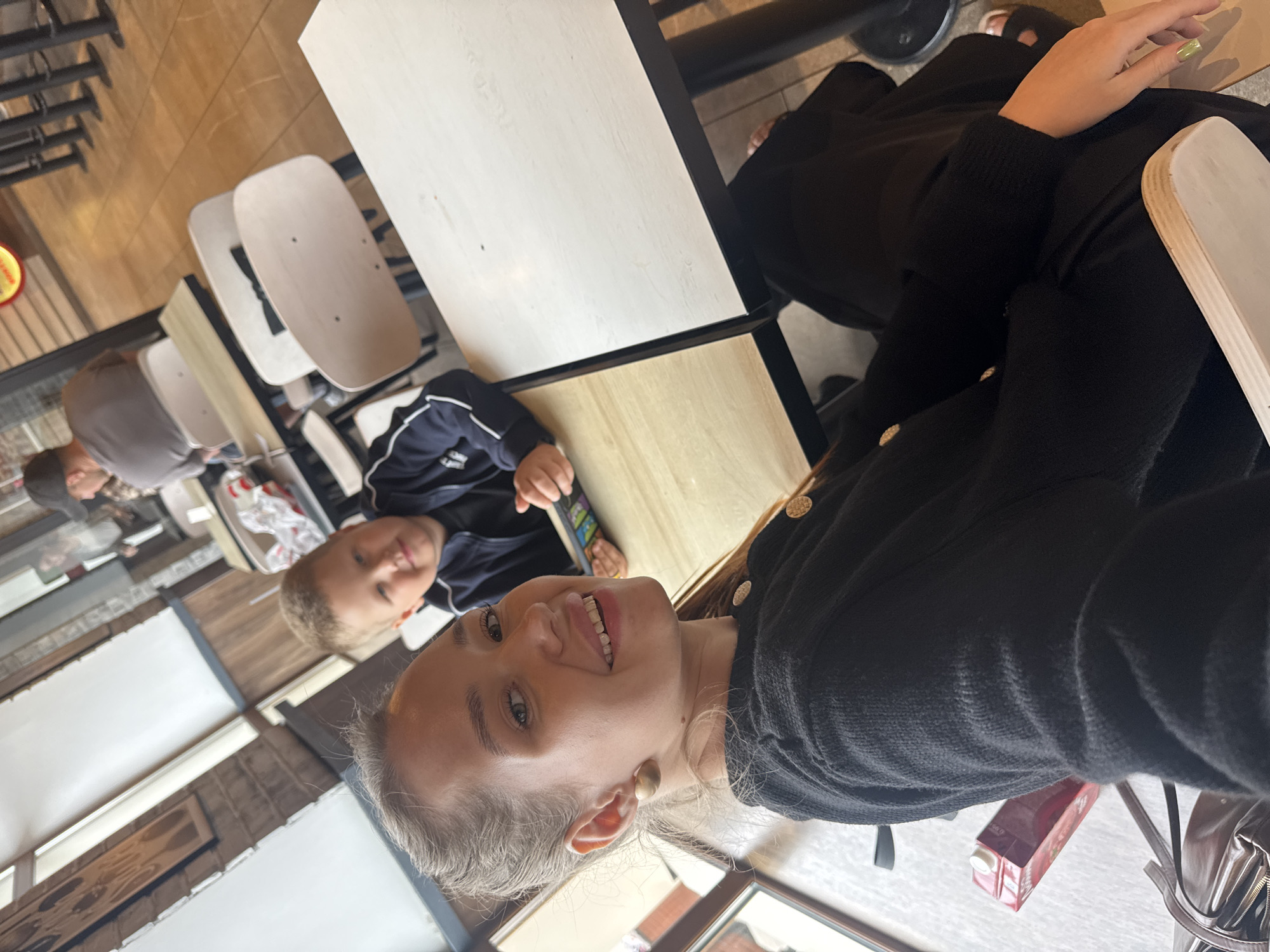 A close-up selfie captures a young woman with blonde hair pulled back, smiling at the camera. She is wearing a black cardigan with gold buttons. To her right and slightly behind her, a young boy with blonde hair, wearing a navy blue jacket with white trim, looks towards the camera with a slight smile. They appear to be sitting at a table in a brightly lit establishment. In the background, other patrons and furniture are visible, suggesting a cafe or restaurant setting. The overall scene is casual and relaxed. The location is in the city of Chaikovsky, Russia.