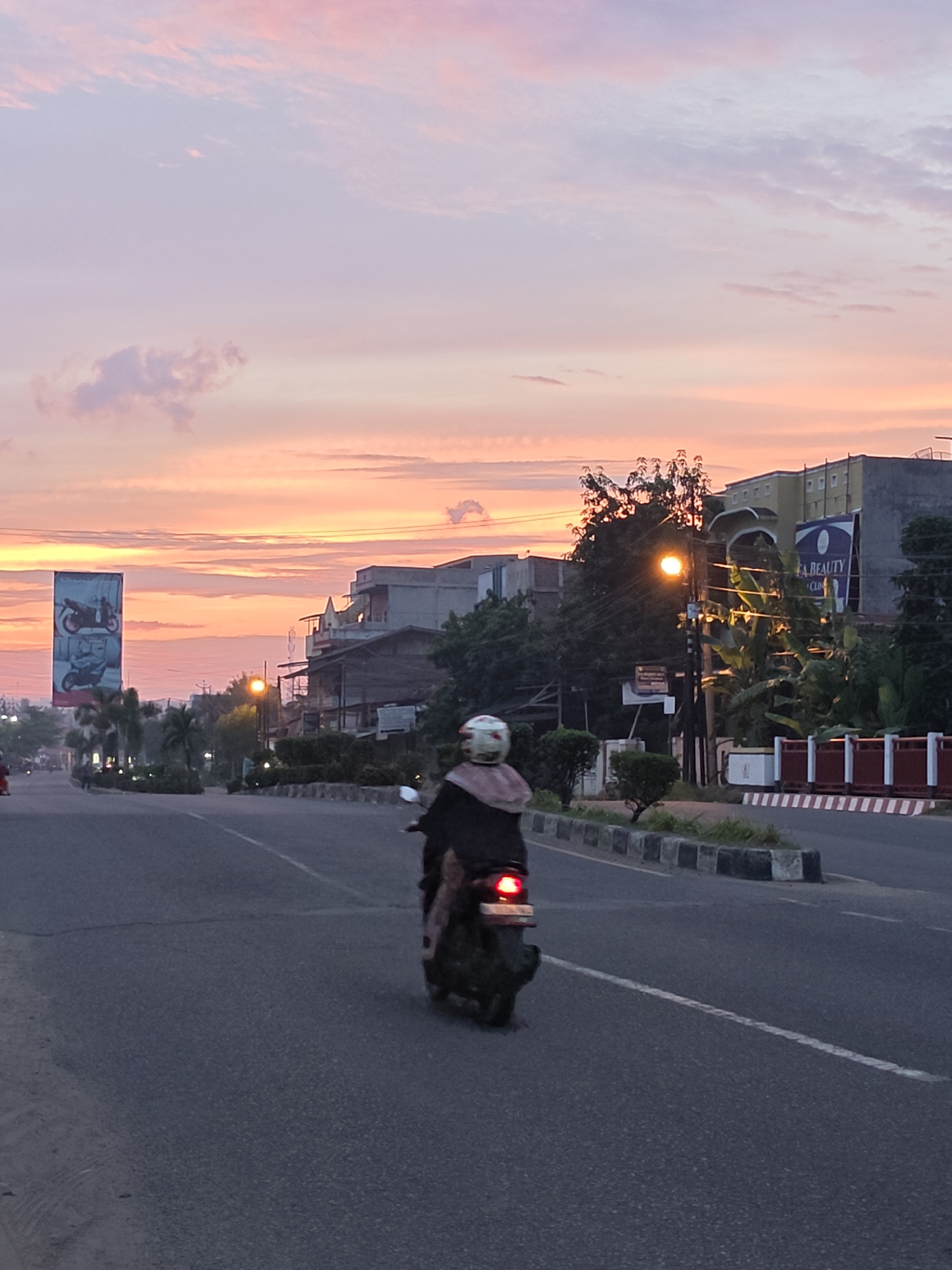 The image captures a street scene in Lhokseumawe, Indonesia, during what appears to be either sunrise or sunset. The sky is dominated by soft hues of pink and orange, with some lighter purple and white clouds. In the foreground, a person is riding a scooter down the asphalt road. The rider is wearing a helmet and a long, dark garment, obscuring their full appearance. The scooter's taillight is illuminated.
On the right side of the road, there are several buildings of varying heights and architectural styles. One building has a prominent sign that reads "A BEAUTY CLINIC." A street lamp with a bright orange light is visible, illuminating the surrounding trees and foliage. The road itself is divided by white lines, and a concrete barrier with white and red stripes separates the road from a strip of greenery and smaller bushes. On the left side of the road, a large vertical billboard displays advertisements for motorcycles. Palm trees and other vegetation are scattered along the roadside, suggesting a tropical or subtropical environment. The overall atmosphere is serene, illuminated by the warm glow of the sky and the street lamp.