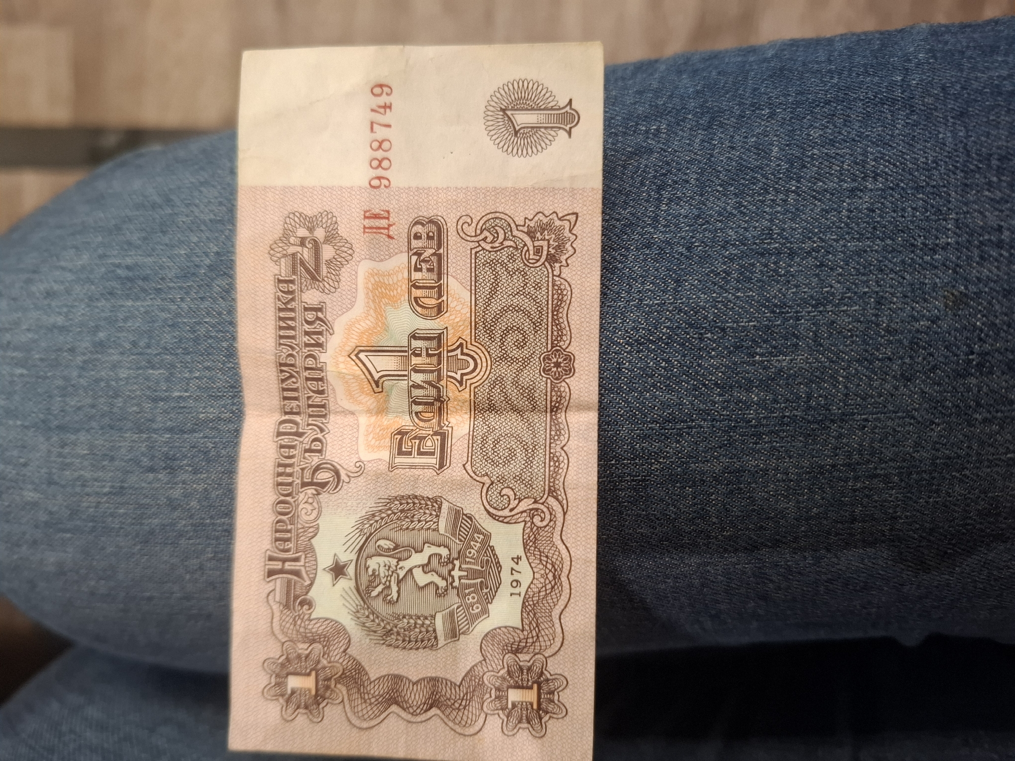 A slightly creased and worn 1 Bulgarian Lev banknote from the People's Republic era is laid vertically on a surface covered by blue denim fabric, likely jeans. The banknote features a faded pinkish-orange and brown color scheme with intricate patterns and designs.
Prominently displayed text on the banknote includes the serial number "ДE 988749" near the top left. Vertically printed on the left side are the words "НАРОДНА РЕПУБЛИКА БЪЛГАРИЯ" (People's Republic of Bulgaria). In the center, also printed vertically, is "1 ЕДИН ЛЕВ" (1 ONE LEV). The numeral "1" also appears in various stylized forms in the top right and bottom corners.
A prominent emblem is located towards the bottom center of the banknote. It depicts a lion rampant within a wreath of wheat ears, topped by a five-pointed star. Below the lion, on a ribbon, are the historical dates "681" and "1944," referring to the foundation of the First Bulgarian Empire and the establishment of the People's Republic. The year "1974" is clearly visible to the right of this emblem, indicating the banknote's issue year.
The background shows the textured blue denim of the clothing, with slight folds, providing a casual context for the object. The lighting is even, suggesting an indoor setting. No specific activity is shown beyond the display of the banknote.