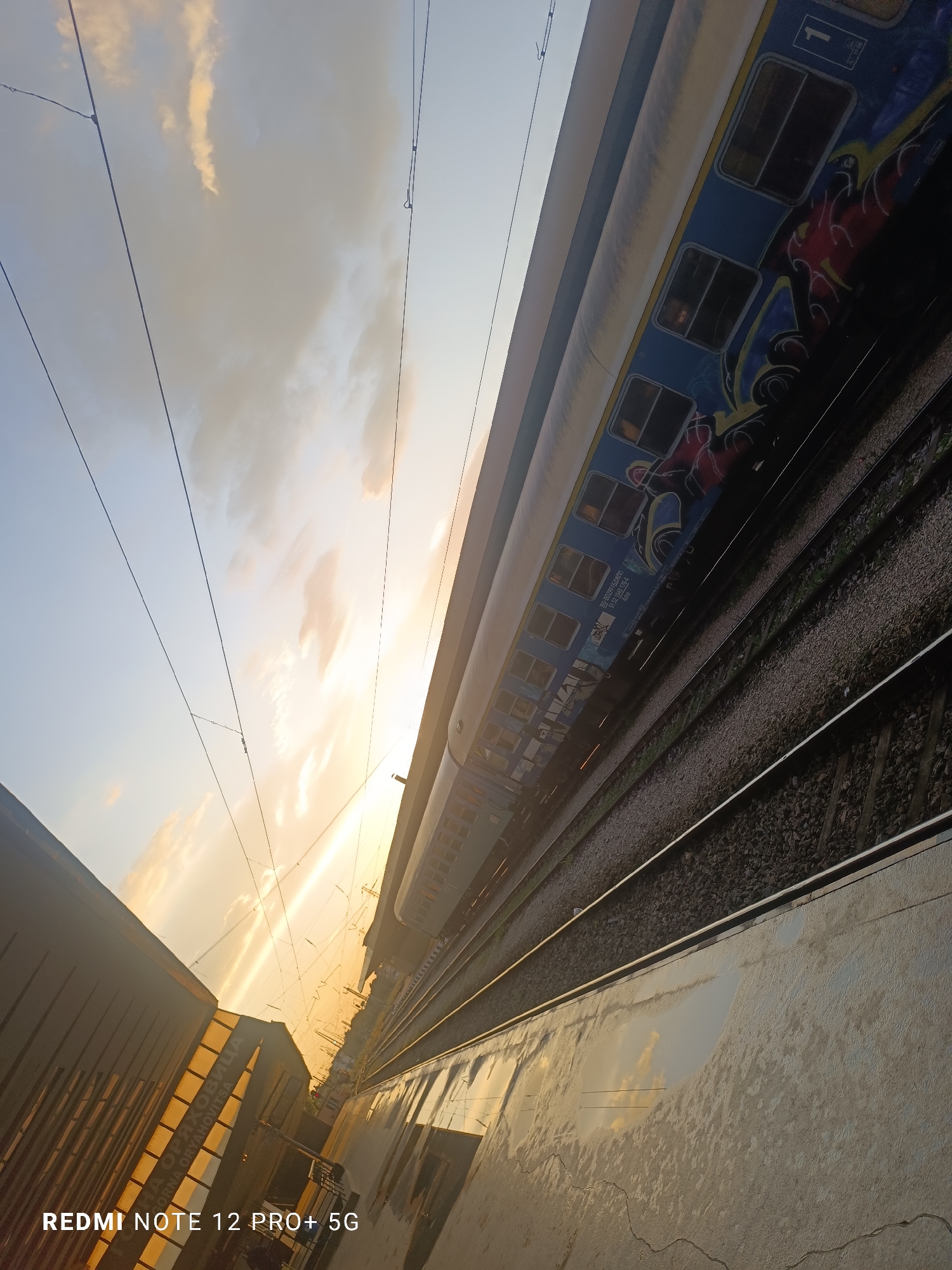 Here is a description of the image:
The photograph presents a ground-level, angled perspective of a train station, likely in Gorna Oryahovitsa, Bulgaria. A portion of a train car occupies the top right, displaying blue and white paint with graffiti-like artwork in red, blue, and yellow. The train tracks run parallel to the platform edge, leading into the distance, where the sun appears to be setting or rising, casting a golden glow across the scene. Overhead wires are visible against the sky.
To the left, a building with a textured facade in shades of orange and brown is seen. The reflective surface of the train platform shows a distorted image of the sky and surrounding architecture. The sky is a gradient of blue and orange, suggesting either sunrise or sunset. "REDMI NOTE 12 PRO+ 5G" is imprinted in the bottom left corner. The train car’s markings include "BG-BOZPP/БДЖПП 51 52 1940 176-4 Ame 1", indicating its association with Bulgarian Railways (BDZ). A sign with Cyrillic script reading "ORNA ORYAHOVITSA" is partially visible on the left building.