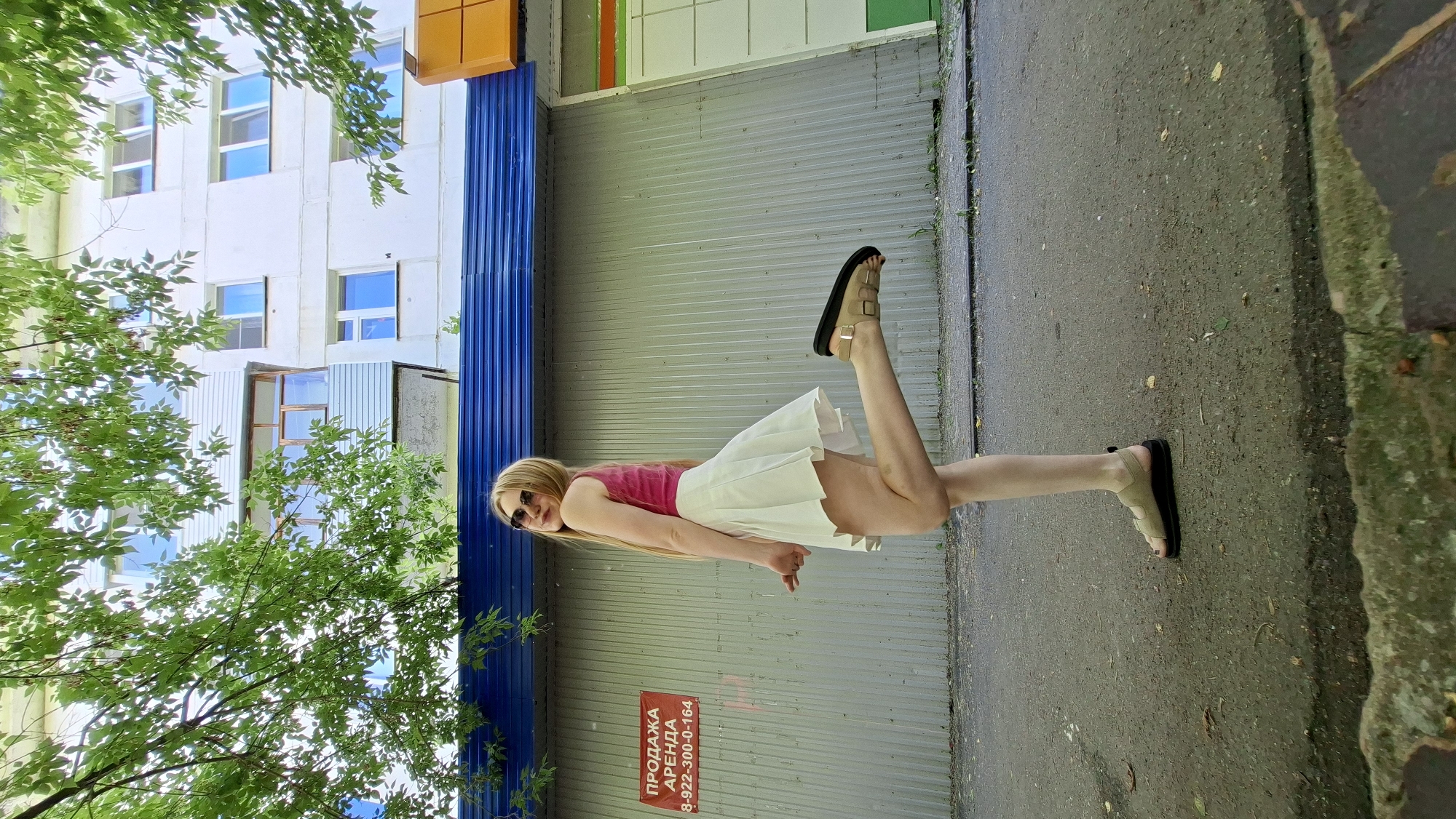 A young woman with long blonde hair and sunglasses is posing outdoors in front of a multi-story building and a corrugated metal wall. She is wearing a pink tank top and a short white pleated skirt, with light brown sandals on her feet. Her right leg is bent at the knee and extended backward. A red sign with Russian text and a phone number is visible on the metal wall behind her. The environment appears to be an urban setting with a paved surface and some greenery from trees. The building is light-colored with multiple windows and balconies. The sky is partially visible through the leaves of trees in the foreground. The image was taken in the city of Chaikovsky, Russia.