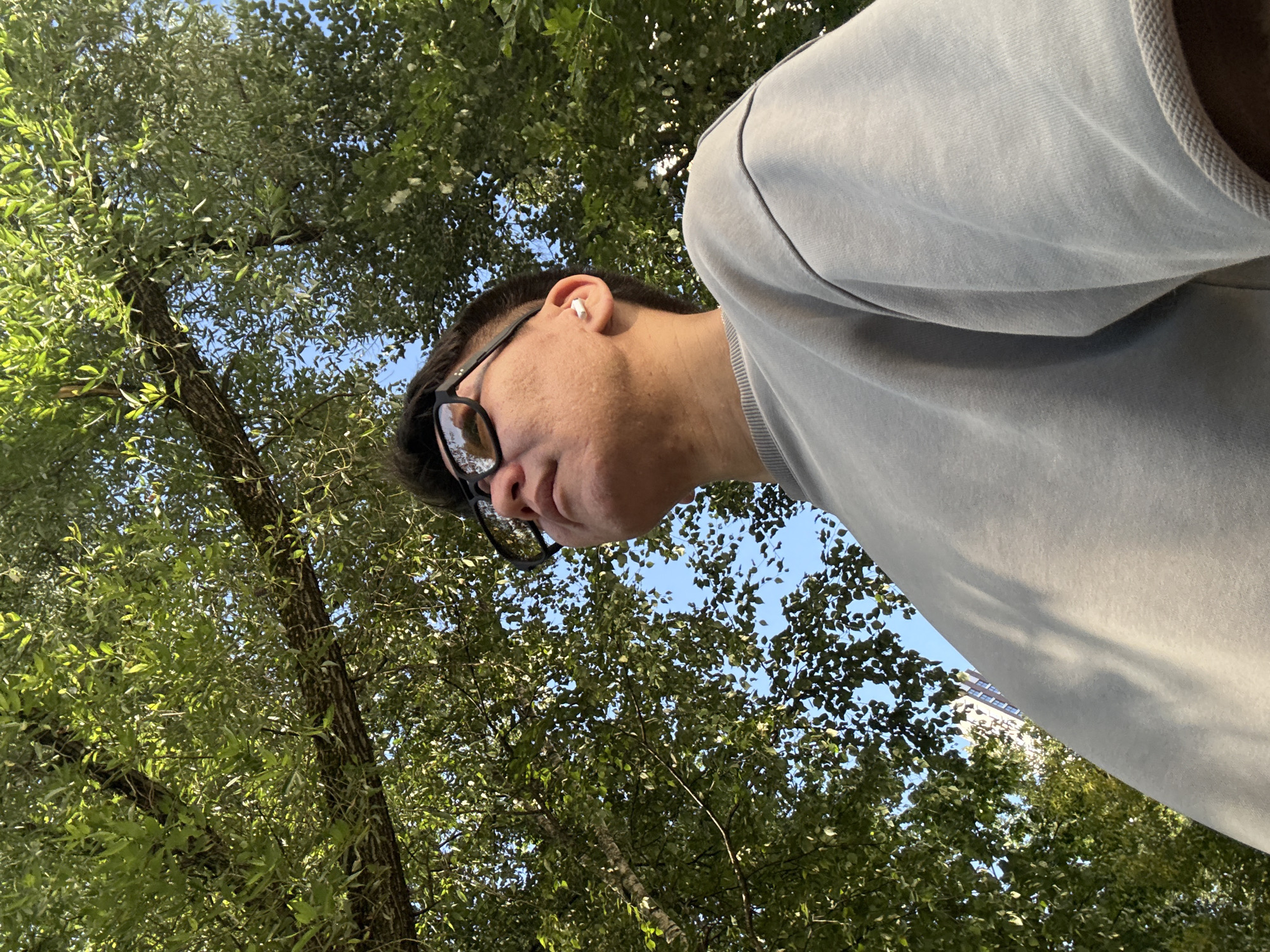 A low-angle, medium shot captures a young person wearing sunglasses and a grey sweatshirt outdoors, looking upwards. They have an earbud in their right ear. The background is dominated by the leafy branches of a tree, with patches of blue sky visible through the foliage. In the distance, a building with several floors can be seen. The overall scene suggests a sunny day in a park or urban green space. The image does not provide any information about the specific location of Moscow, Russia.