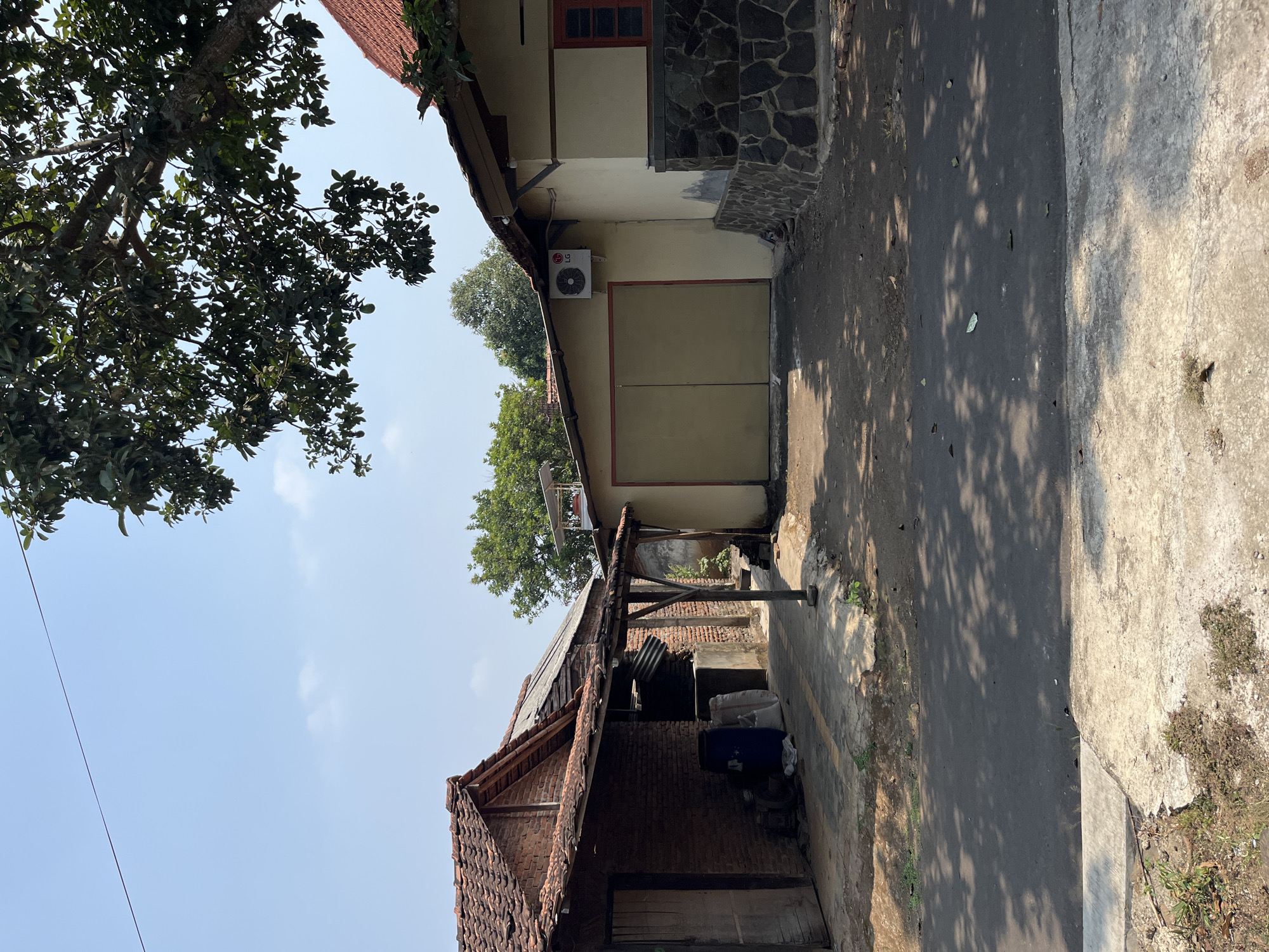 The image depicts a residential scene in Glagah Kulon, Indonesia, under a clear blue sky with scattered clouds. In the foreground, an asphalt road is visible, shaded by dappled sunlight filtering through trees. To the left, a house with a red tile roof and brick walls is partially obscured by an overhang. This structure appears older, with some visible damage to its roof. Adjacent to it, a newer, lighter-colored building with a red tile roof and a stone-clad lower section is seen. This building features a large, closed, light-colored garage door and an outdoor air conditioning unit with an LG logo mounted on the wall. The overall environment suggests a humble, possibly rural or suburban, dwelling with a mix of older and newer constructions. No people are present in the image.