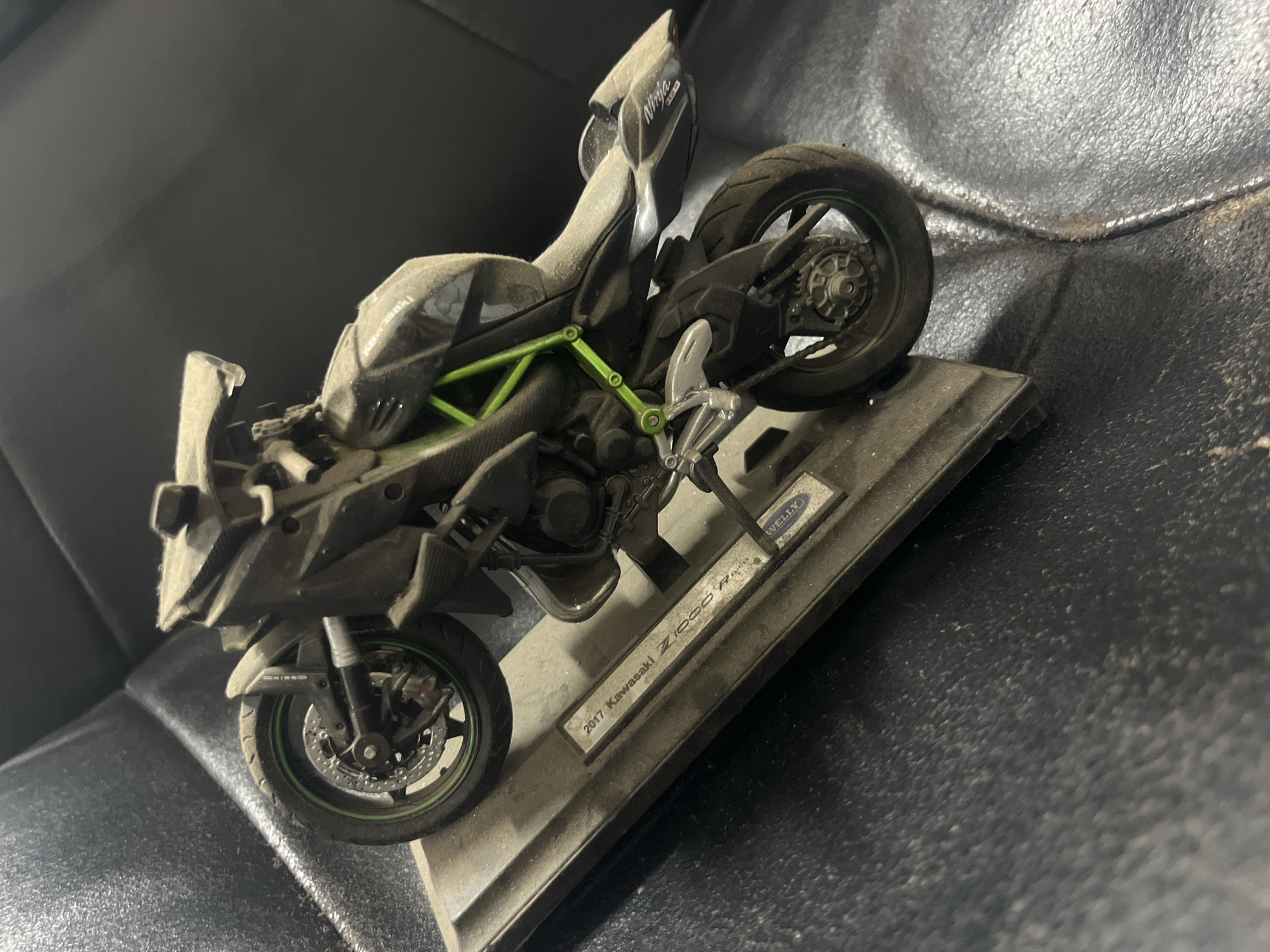 A dusty, 1:12 scale model of a 2017 Kawasaki Z1000 R Edition motorcycle, manufactured by Welly, is displayed on a black leather surface. The motorcycle is dark gray with bright green frame accents and green pinstripes on the wheels. The model is positioned on a rectangular base with the model's identification printed on a metal plate. The background is out of focus and appears to be a dark, textured surface. The lighting suggests an indoor setting, possibly a home or a collector's display. The image is taken from a medium close-up, slightly angled view, focusing on the motorcycle and its display base. The location in Burgas, Bulgaria, is not determinable from the image.