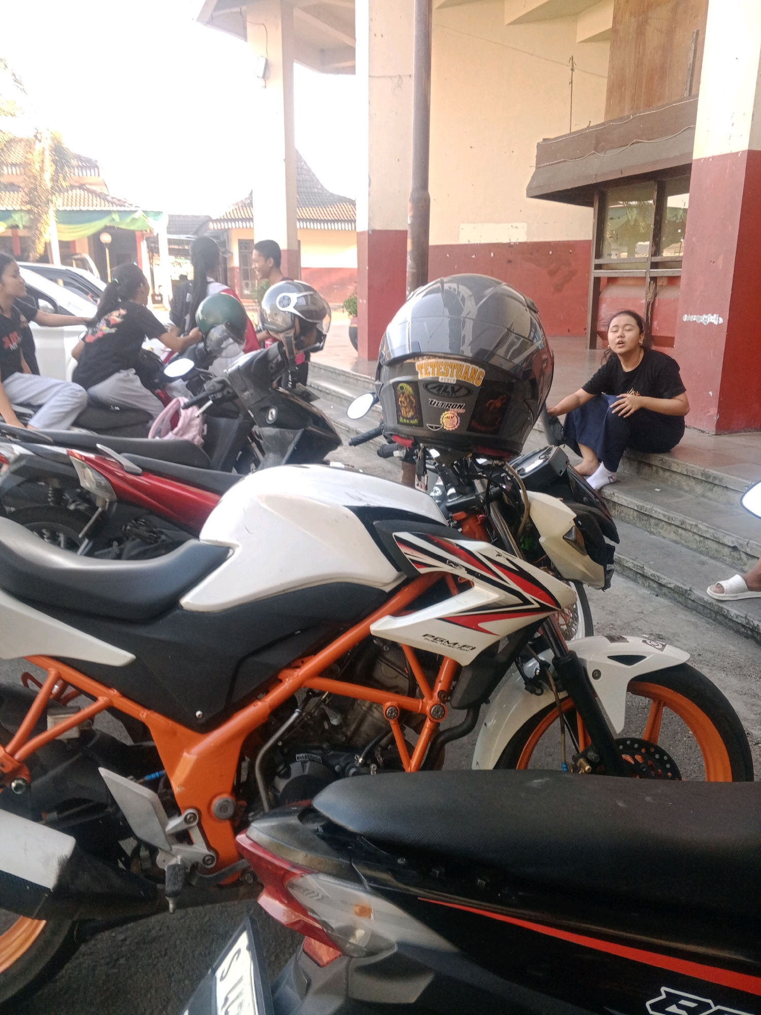 In Wuryorejo, Indonesia, several individuals are gathered near parked motorcycles under a building with red and cream-colored walls. In the foreground, a white and orange motorcycle is prominently displayed, featuring an orange frame and wheels. A dark helmet with stickers is placed on its handlebars. To the right, a young woman wearing a black t-shirt and dark skirt is seated on concrete steps, looking upwards and appearing to speak. Behind her, other individuals, including a woman in a black t-shirt and patterned pants, and a person in a red shirt, are seen near other motorcycles and a white car. The scene appears to be an outdoor area adjacent to a building, possibly a school or community center, with sunlight casting shadows.