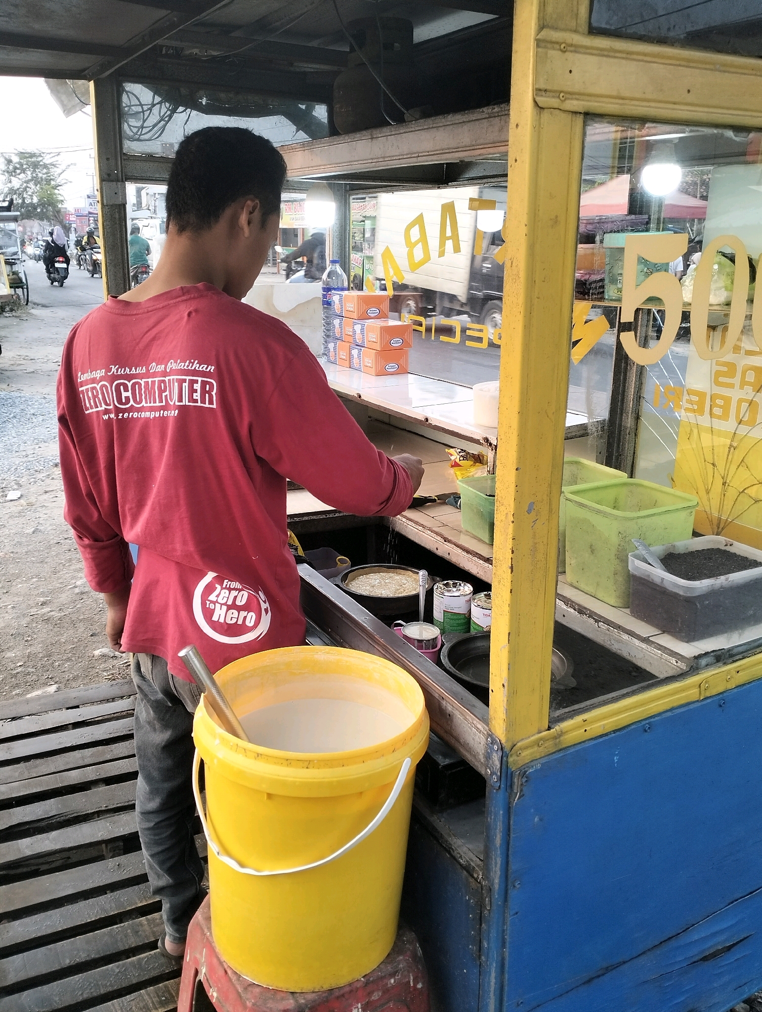 A person wearing a red long-sleeved shirt and dark pants stands behind a food cart in Perumnas Way Kandis, Indonesia. The shirt has white lettering on the back that reads "Lembaga Kursus Dan Pelatihan ZERO COMPUTER" and a circular logo with "From Zero To Hero". A large yellow bucket containing a light-colored liquid and a metal scoop is placed on a red stool to the left of the person. The food cart has a blue front panel, a yellow frame around the glass display, and a yellow top. Inside the cart, there are various containers holding ingredients, including two green plastic tubs, one dark-colored tub with a spoon, and a pan with food. Boxes are stacked on a shelf inside the cart. Through the glass, a truck with "B A" visible on its side is seen, along with a street scene with motorbikes and pedestrians. The ground around the cart is paved with wooden planks.
