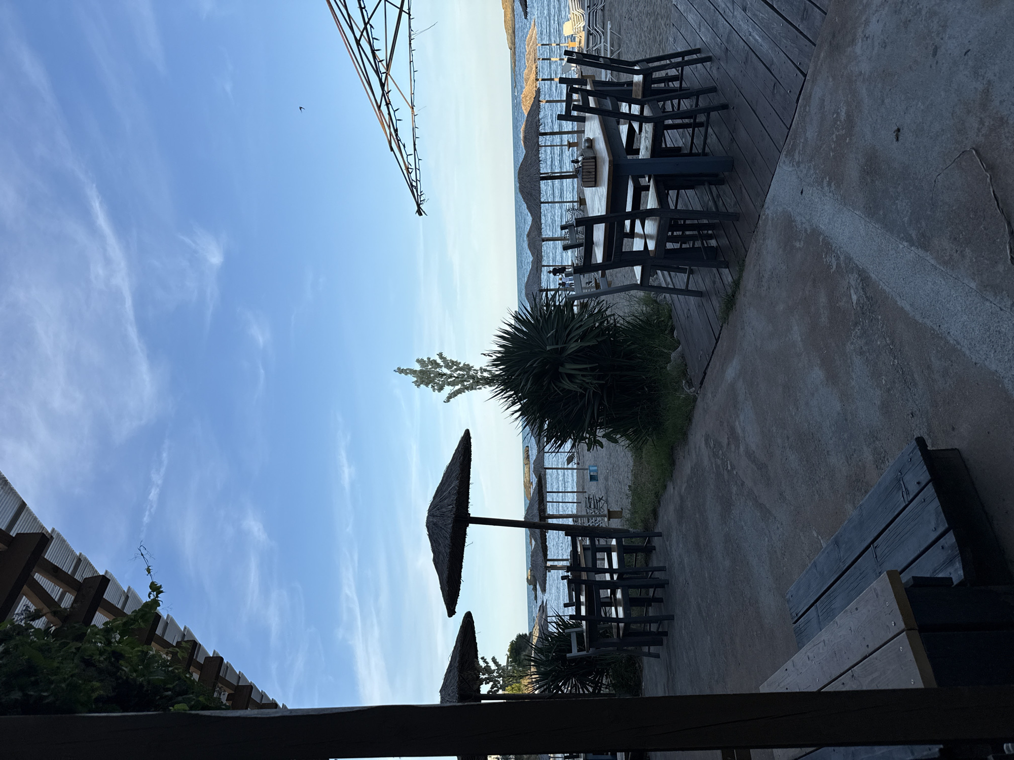 The image depicts an outdoor seating area at a beachside establishment in Tsarevo, Bulgaria. In the foreground, a dark wooden picnic bench is visible on a concrete surface. To the right, a wooden deck leads to tables and chairs set up on the sand, facing the Black Sea. Several straw umbrellas provide shade over the beach, with lounge chairs arranged further back. The water is calm, and the sky is a clear blue with scattered wispy clouds. A lone bird is silhouetted against the sky in the upper right quadrant. The overall scene suggests a relaxed atmosphere at dusk or early evening.