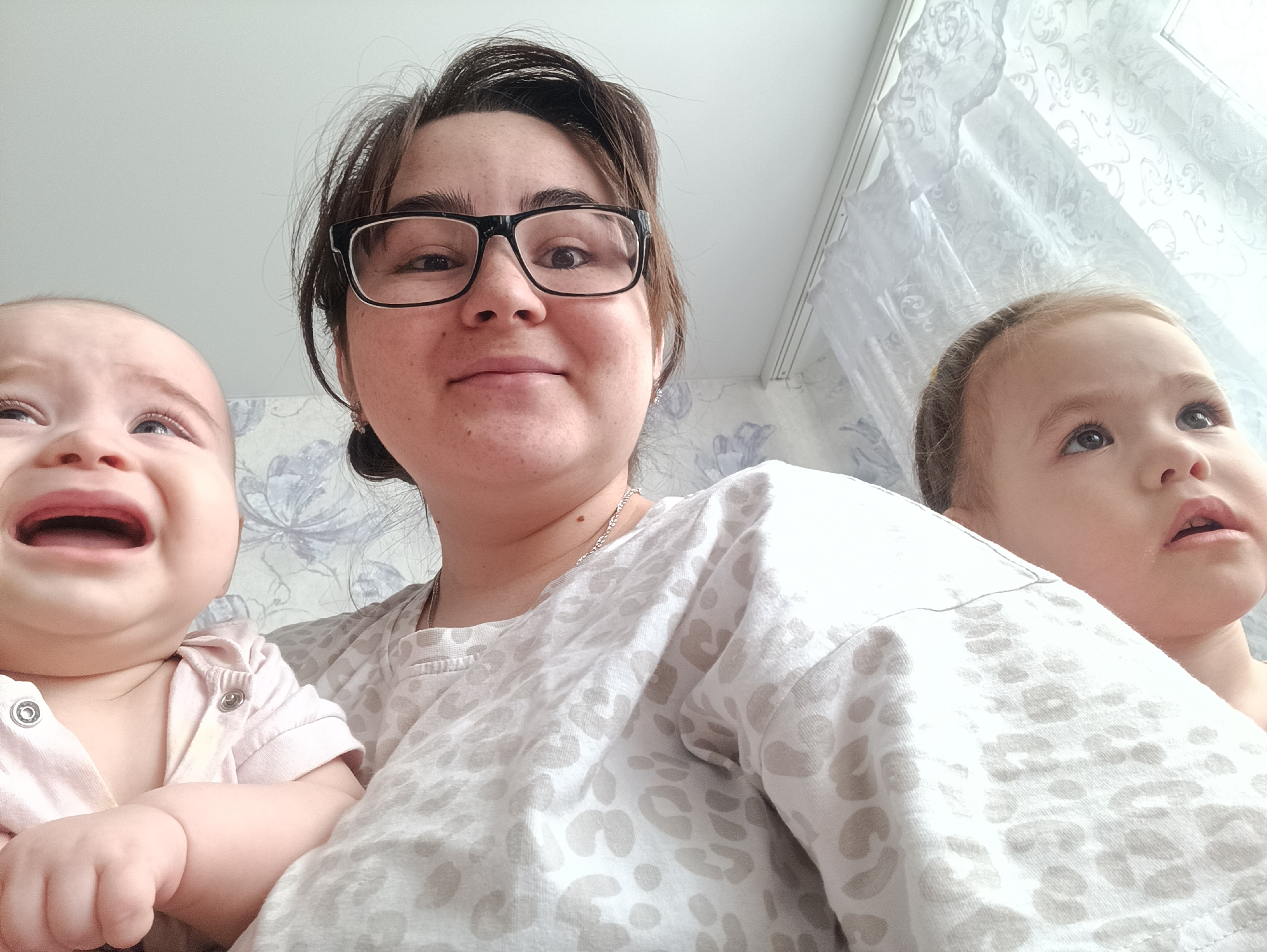 The photograph presents a selfie-style composition featuring a woman, likely in her late 20s or early 30s, positioned centrally and holding two young children. The woman is wearing glasses and a light-colored, patterned top. To her left, a baby is crying, its mouth open in distress, with tears visible. To her right, a slightly older child looks upwards with an inquisitive expression. The background suggests an indoor setting, possibly a domestic environment, with a patterned wall and a window covered with a white lace curtain. Based on available information, this photograph was taken in Noviy, Russia.